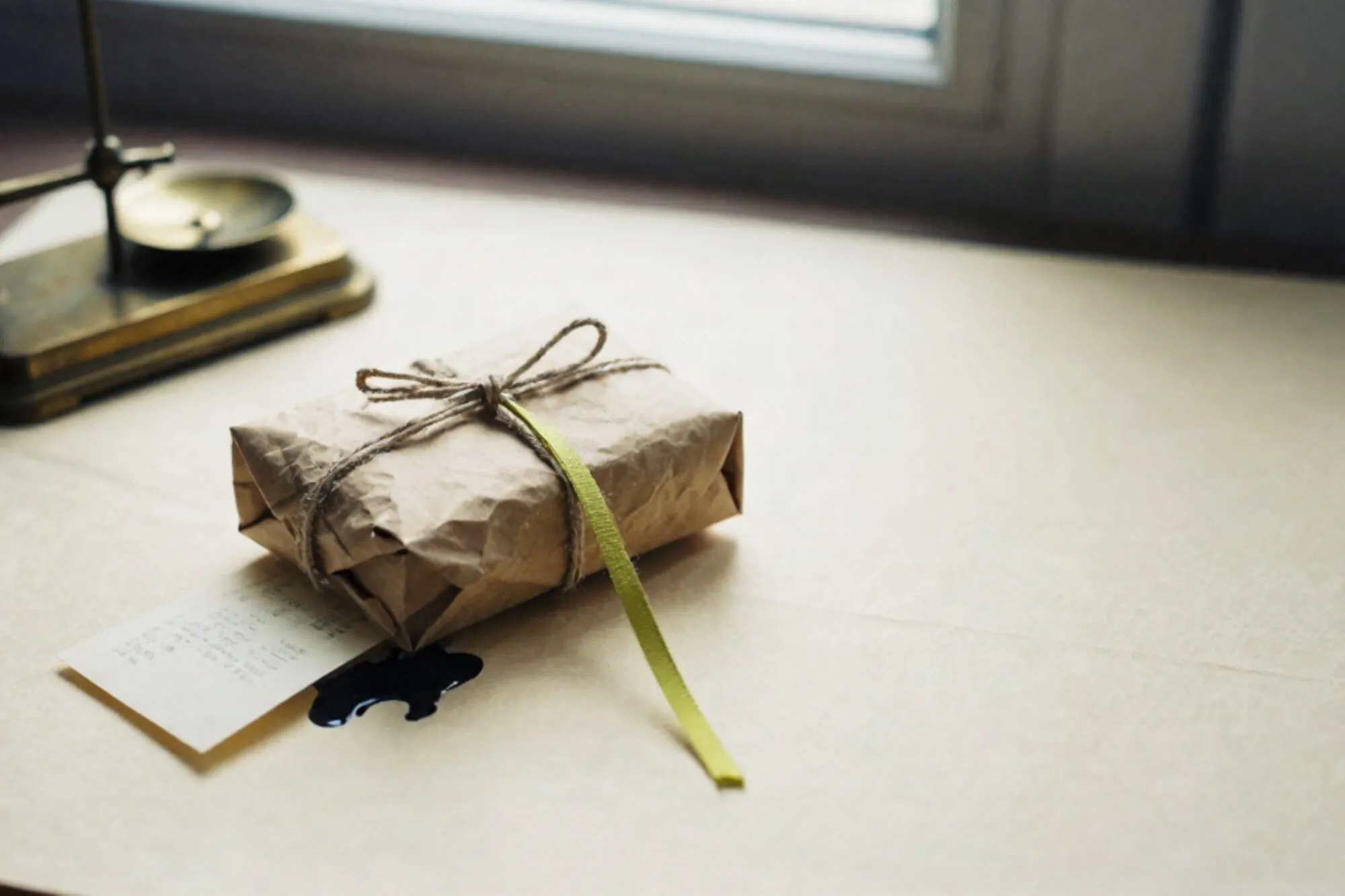 Craft-paper parcel with linen twine and green ribbon, brass scale, folded receipt on ivory paper surface.