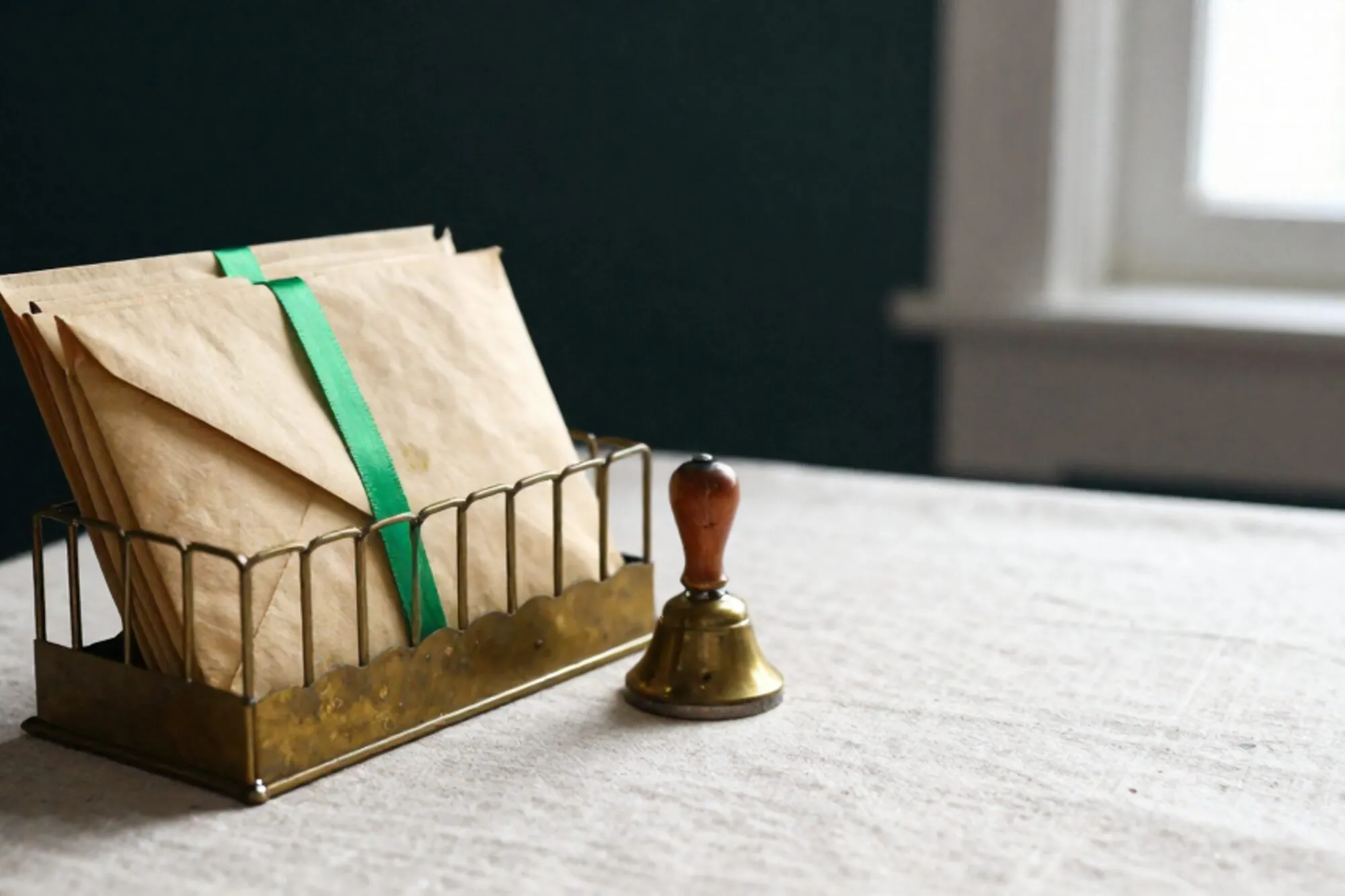 Brass mail sorting rack on ivory linen with manila envelopes, one tied in chartreuse ribbon, brass bell, date stamp.