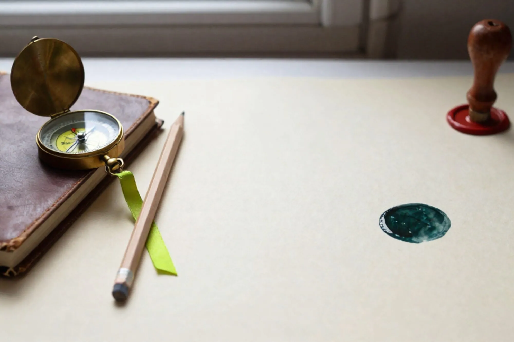 Brass compass, leather notebook, cedar pencil, green ribbon, red wax seal on ivory paper by a window.