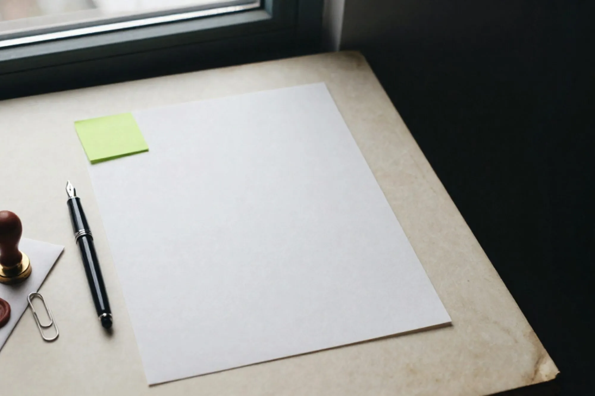 Worksheet on ivory desk, green sticky note, fountain pen, brass paper clip, wax seal on envelope, side window light.