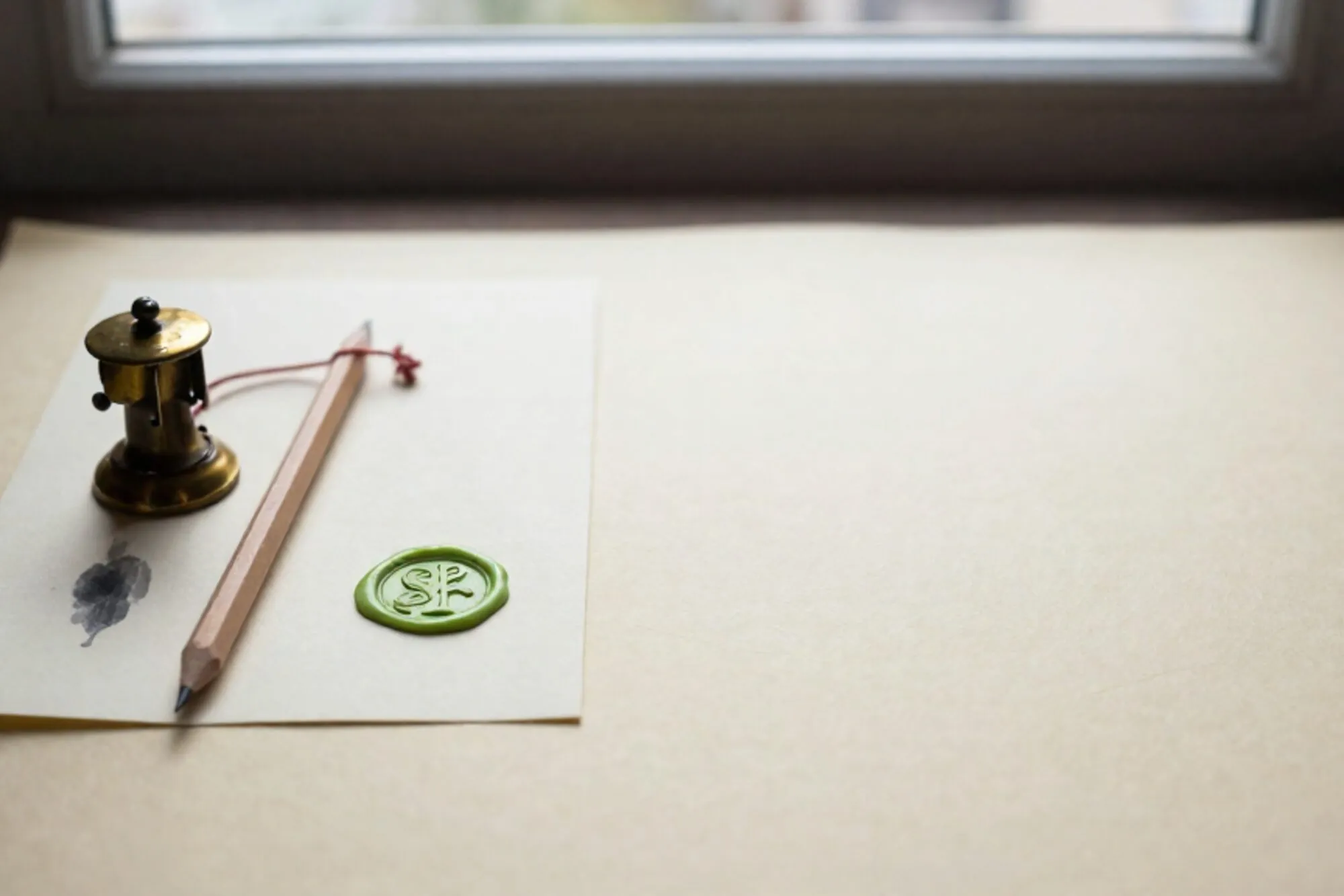 Brass composing stick, pencil with red thread, folded paper with green wax seal on ivory surface.