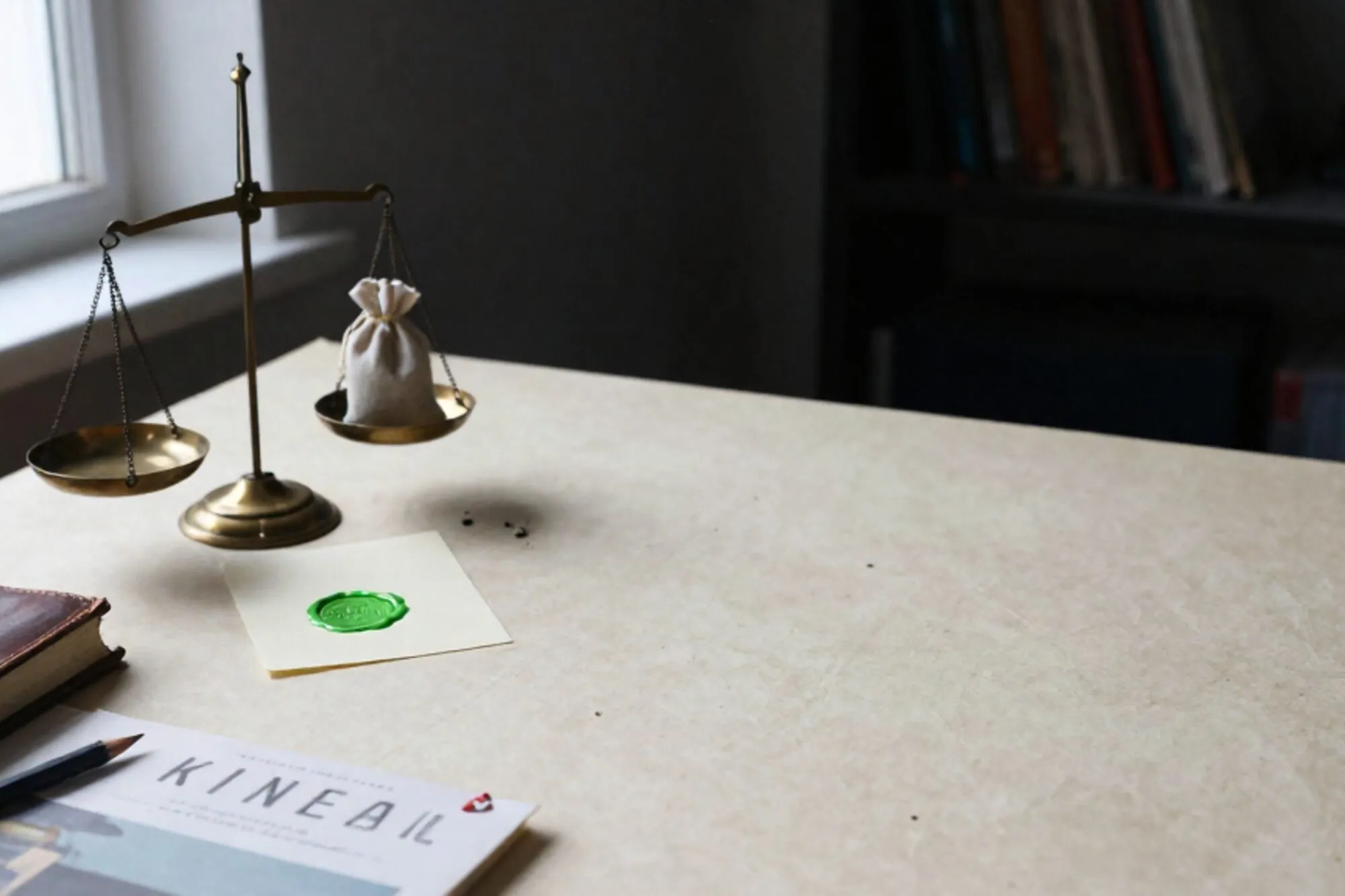 Brass balance scale weighing two linen pouches, cream card with green wax seal, leather ledger, pencil on ivory desk.