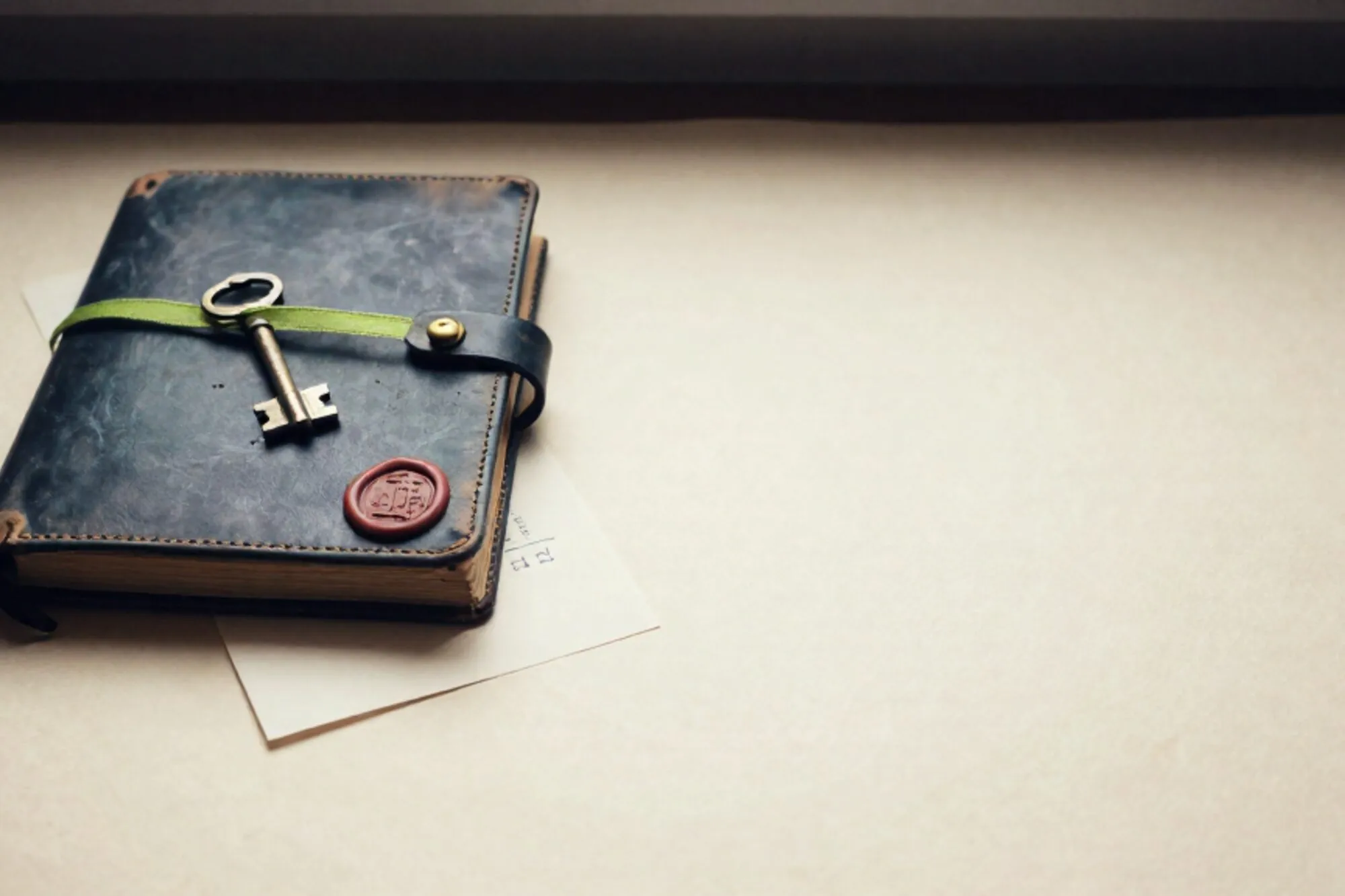 Leather ledger with brass clasp, brass key, green ribbon, red wax seal on cream card, ivory paper surface.