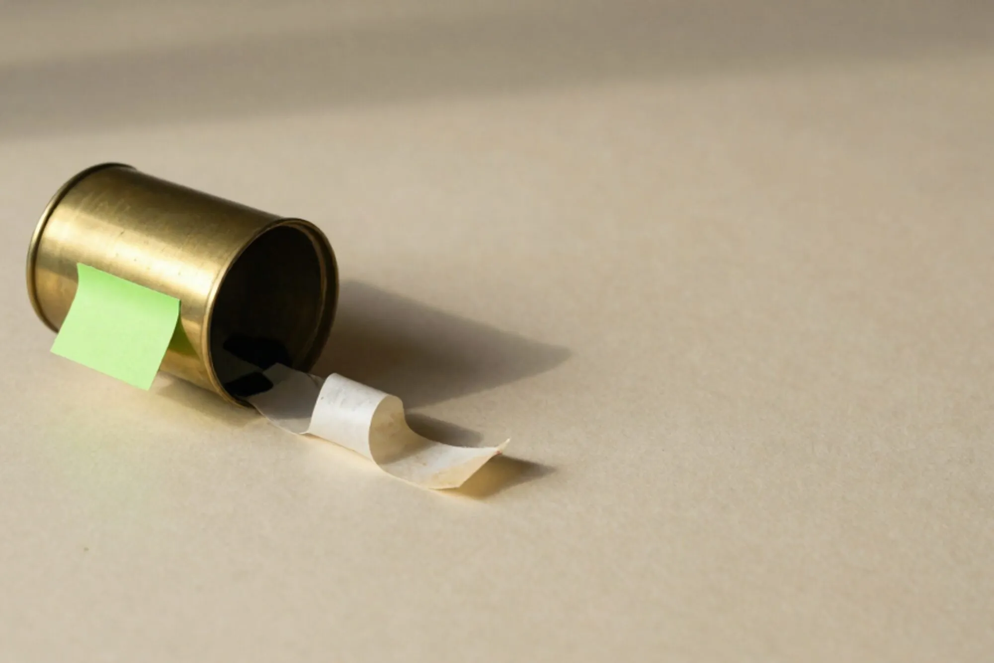 Brass pneumatic tube canister tipped open on ivory paper, receipt tape uncoiling, chartreuse sticky note on its side.