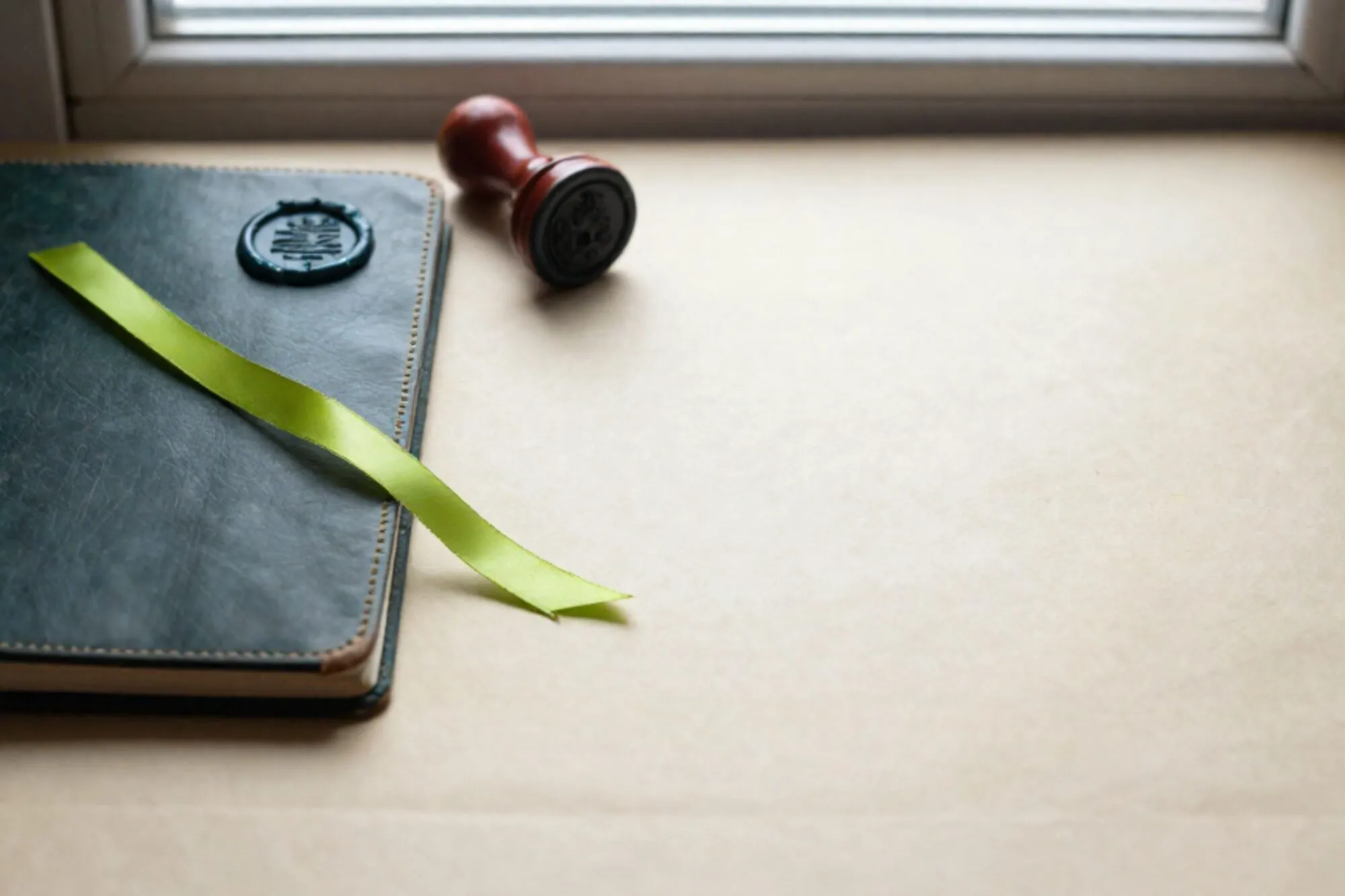 Closed leather ledger with green silk ribbon, wax seal and clay-red stamp on ivory paper by a window.
