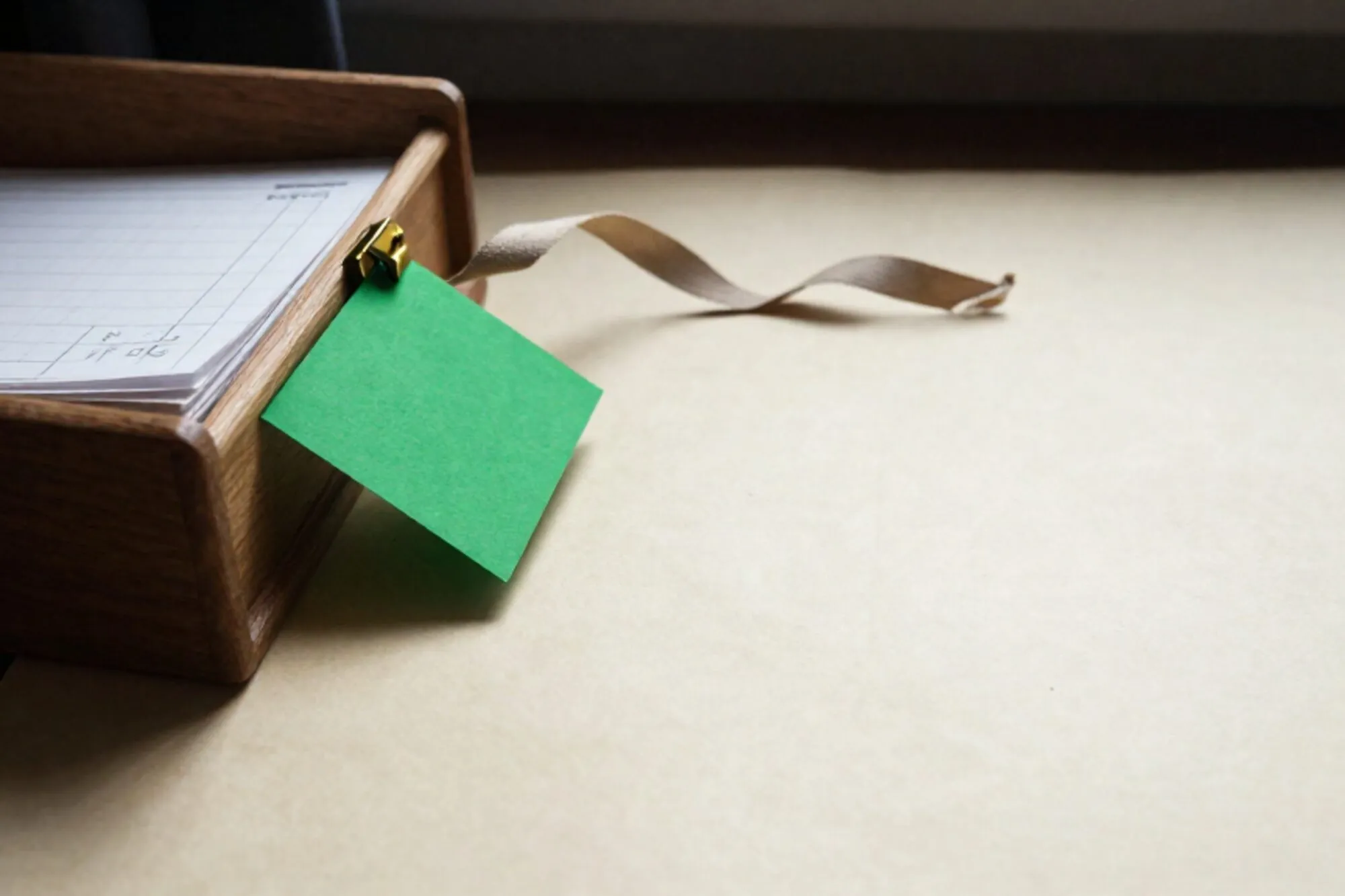 Open eiken kaartenbak met grootboekpapier, koperen tussenschot, één groene kaart en linnen lint op een vloeiblad bij raam.
