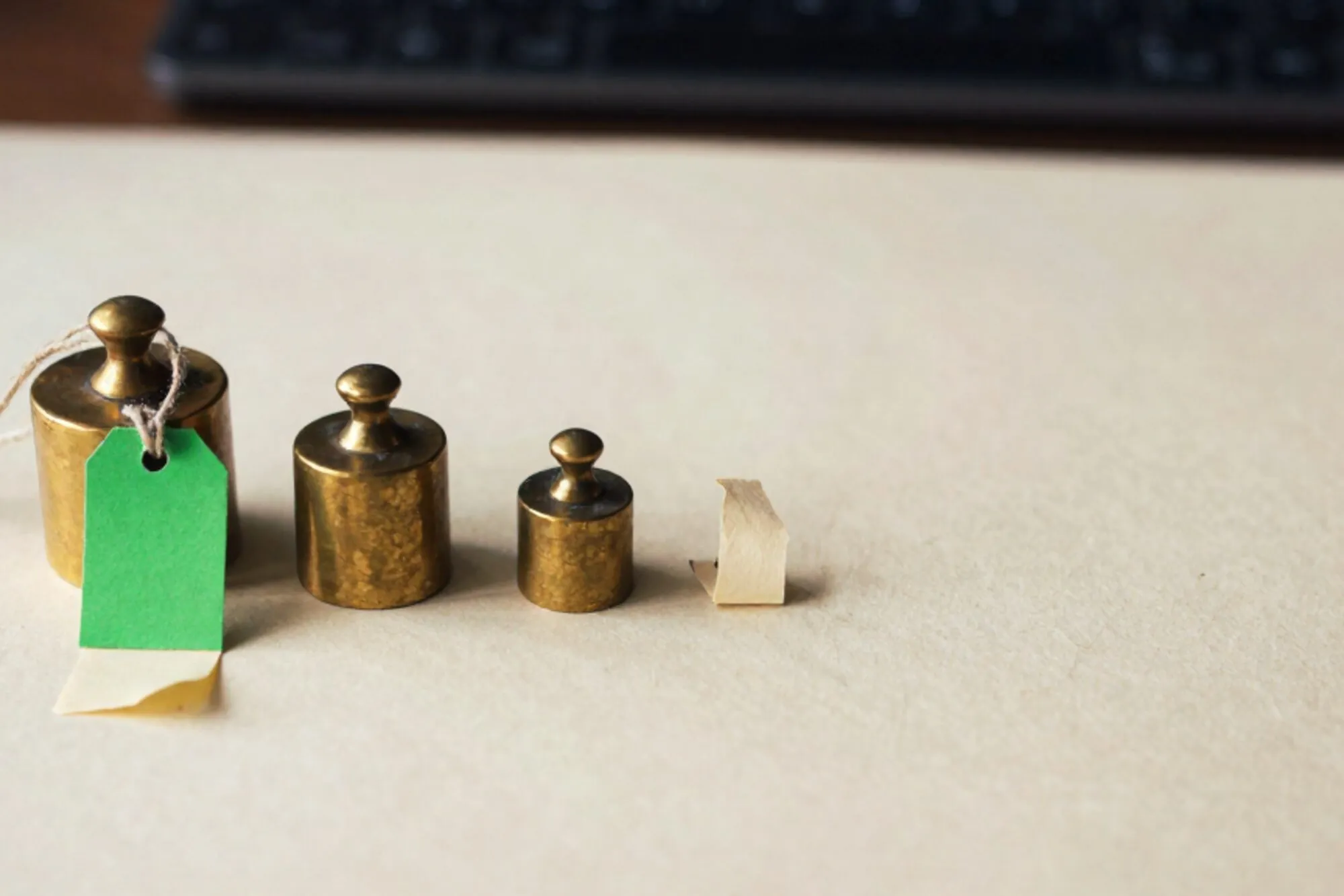 Three brass apothecary weights in a row, green price tag on twine, curled cream receipt, ivory paper surface, side light.