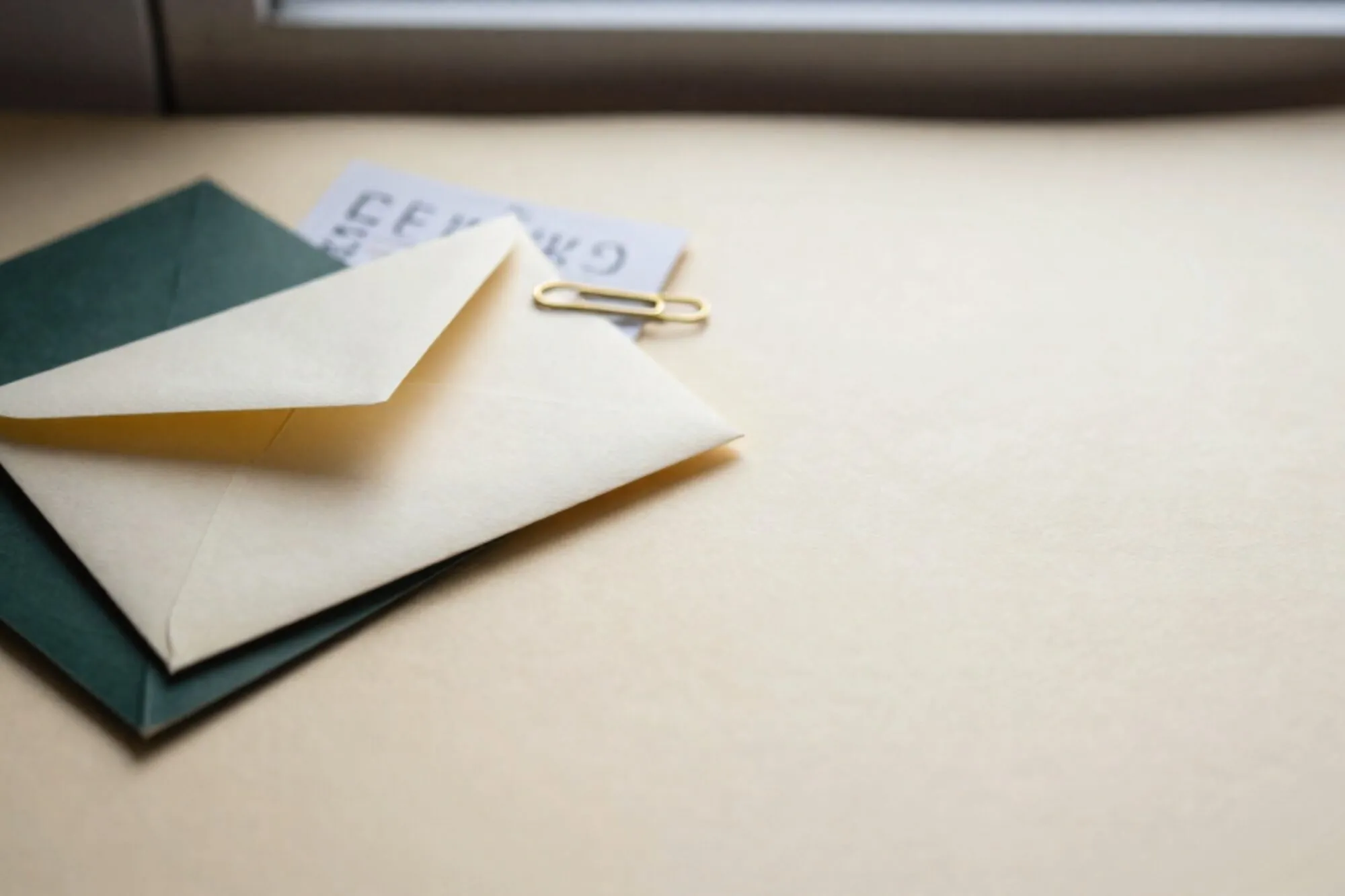Crème envelop met limegroen lint op donkergroen vloeiblad, koperen paperclip en gevouwen bon, ivoorkleurig papier.