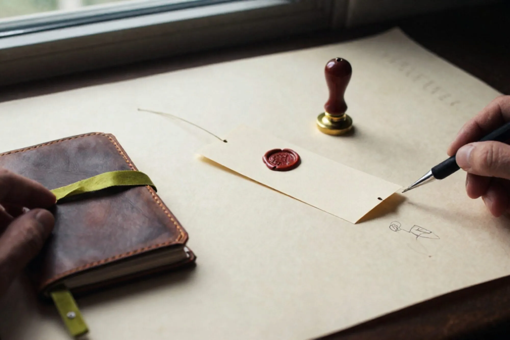 Paper ticket on brass spike, leather notebook with green ribbon, red wax seal on ivory paper surface.