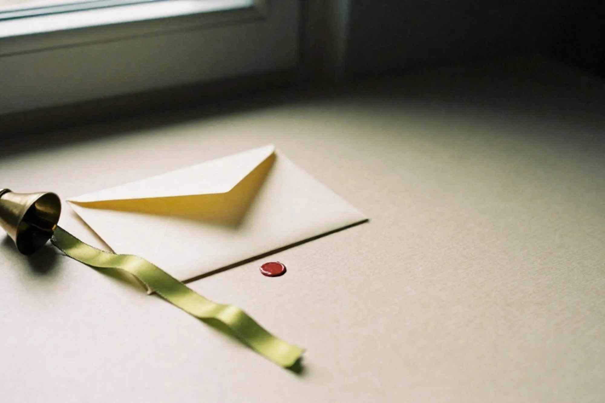 Cream envelope unsealed on ivory blotter, chartreuse silk ribbon, tipped brass bell, broken clay red wax seal.
