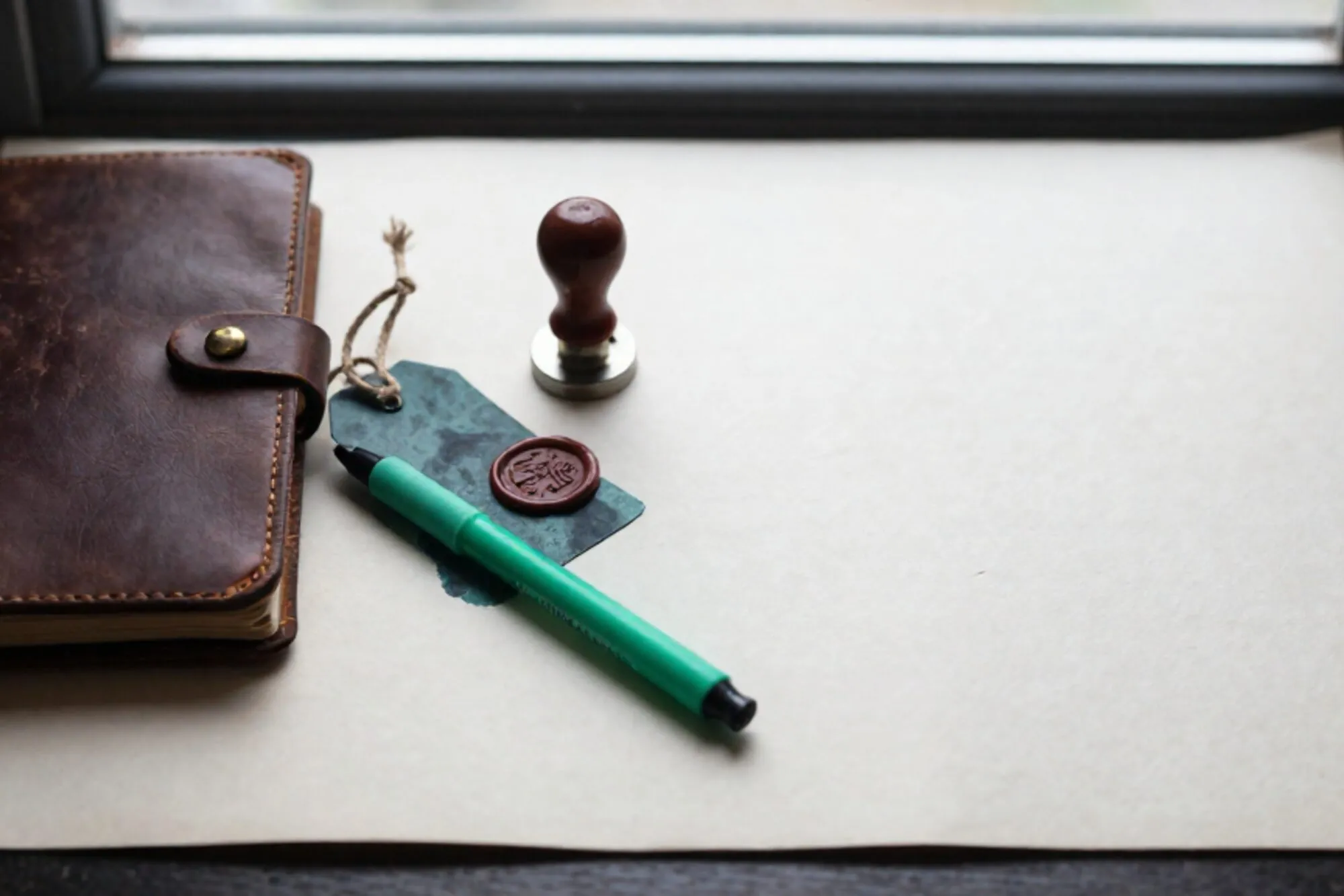 Closed leather logbook with brass clasp, iron shipping tag on linen twine, green ribbon marker, red wax seal on ivory paper.