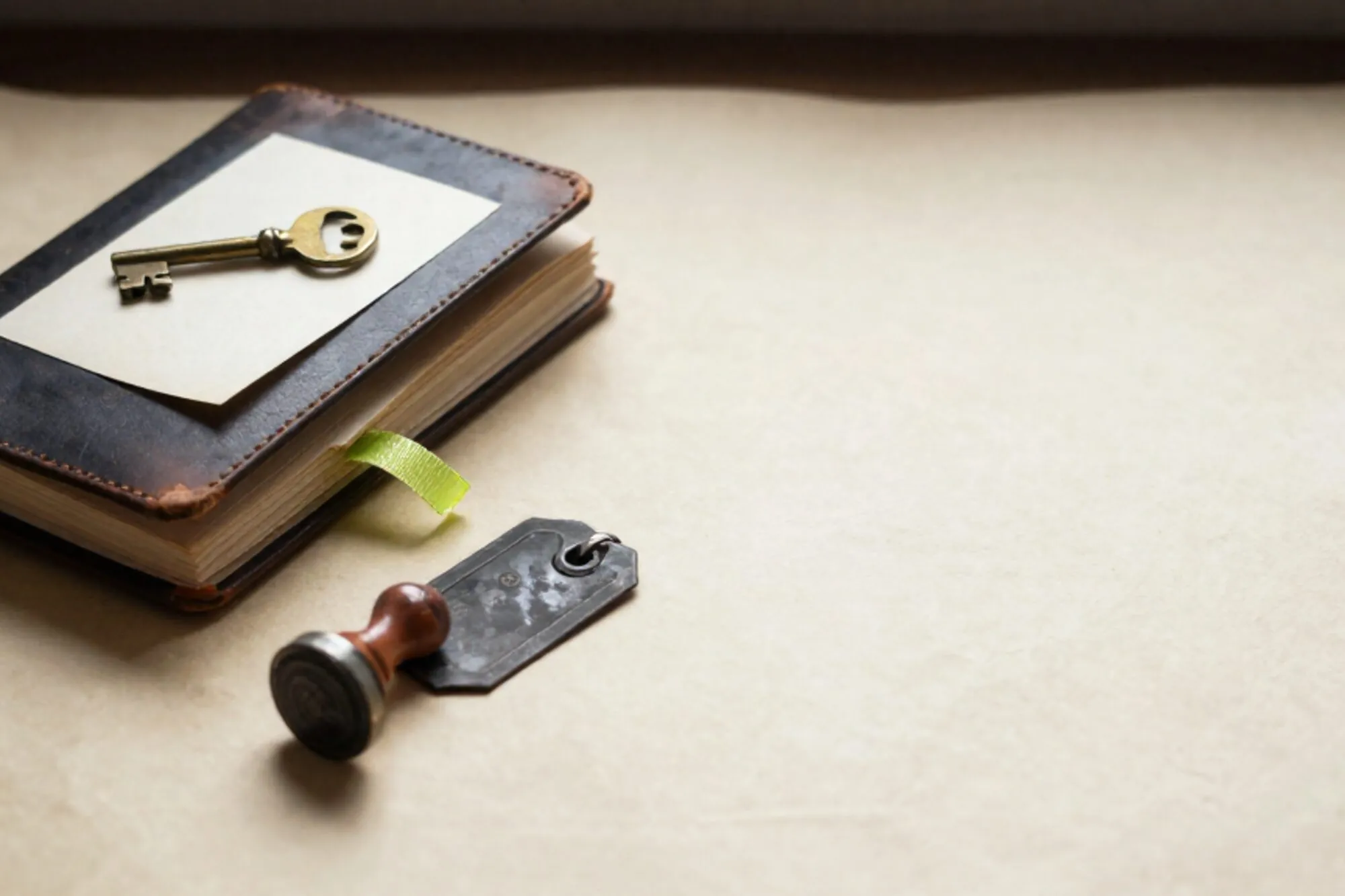 Gesloten leren logboek met messing sleutel op cream kaart, groen lint, ijzeren label en rubberen stempel op ivoor linnen.