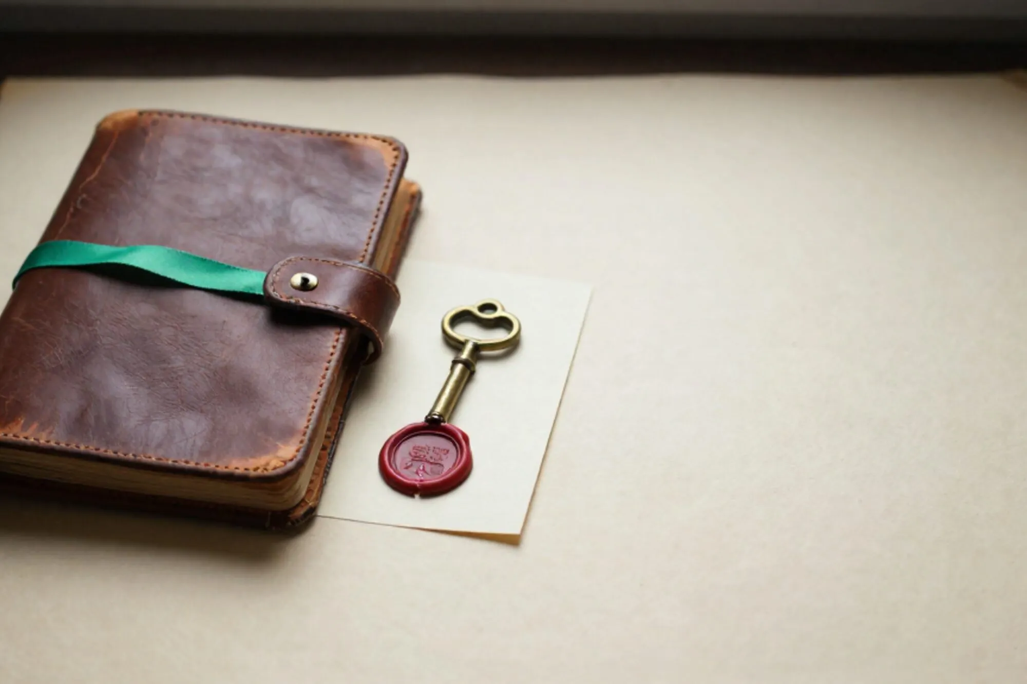 Open leather logbook with green ribbon, tarnished brass key on cream card, cracked red wax seal on ivory paper.