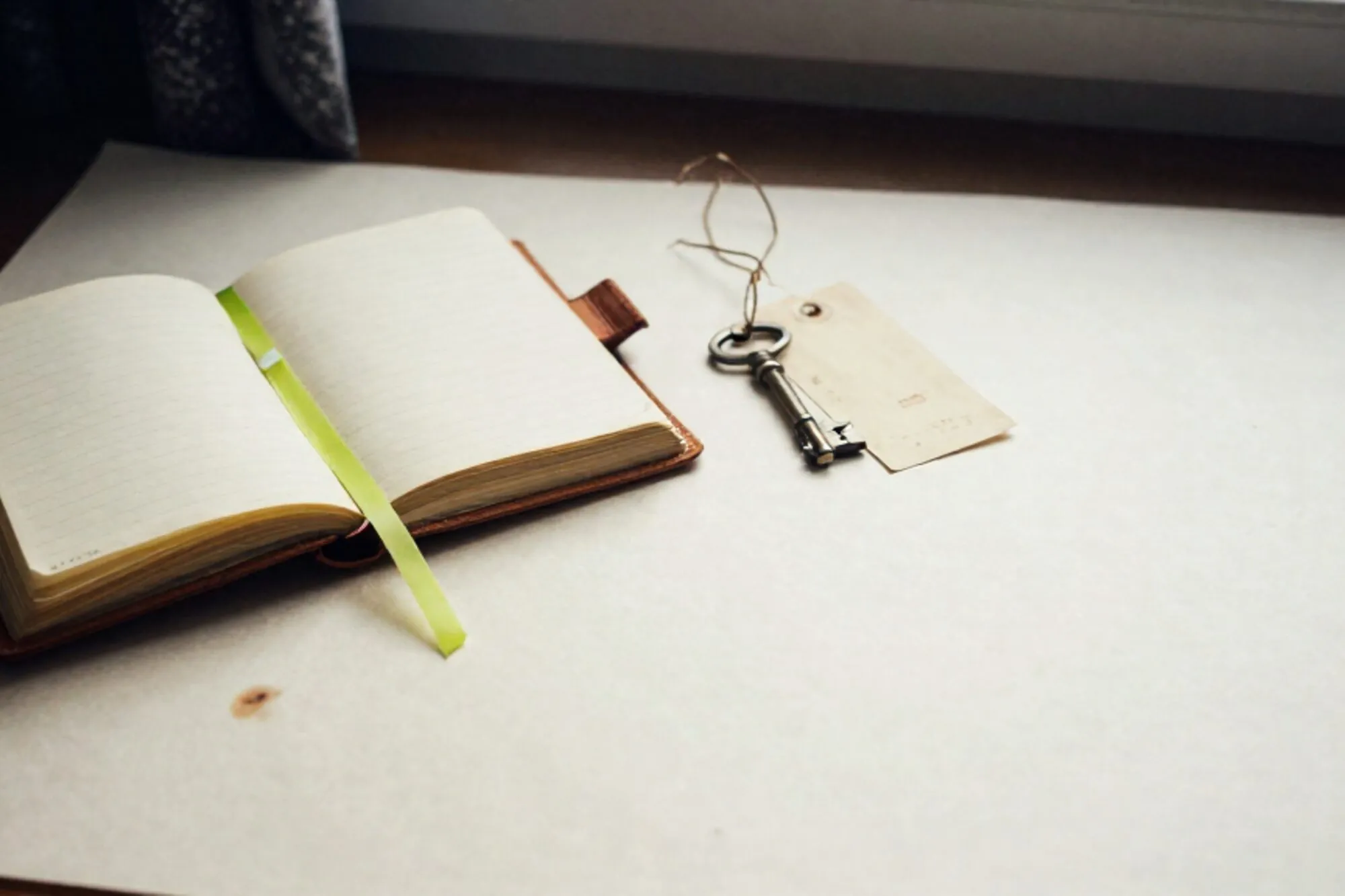 Open leather ledger with paper tabs and green ribbon, brass key on index card, twine shipping tag, ivory paper, side light.