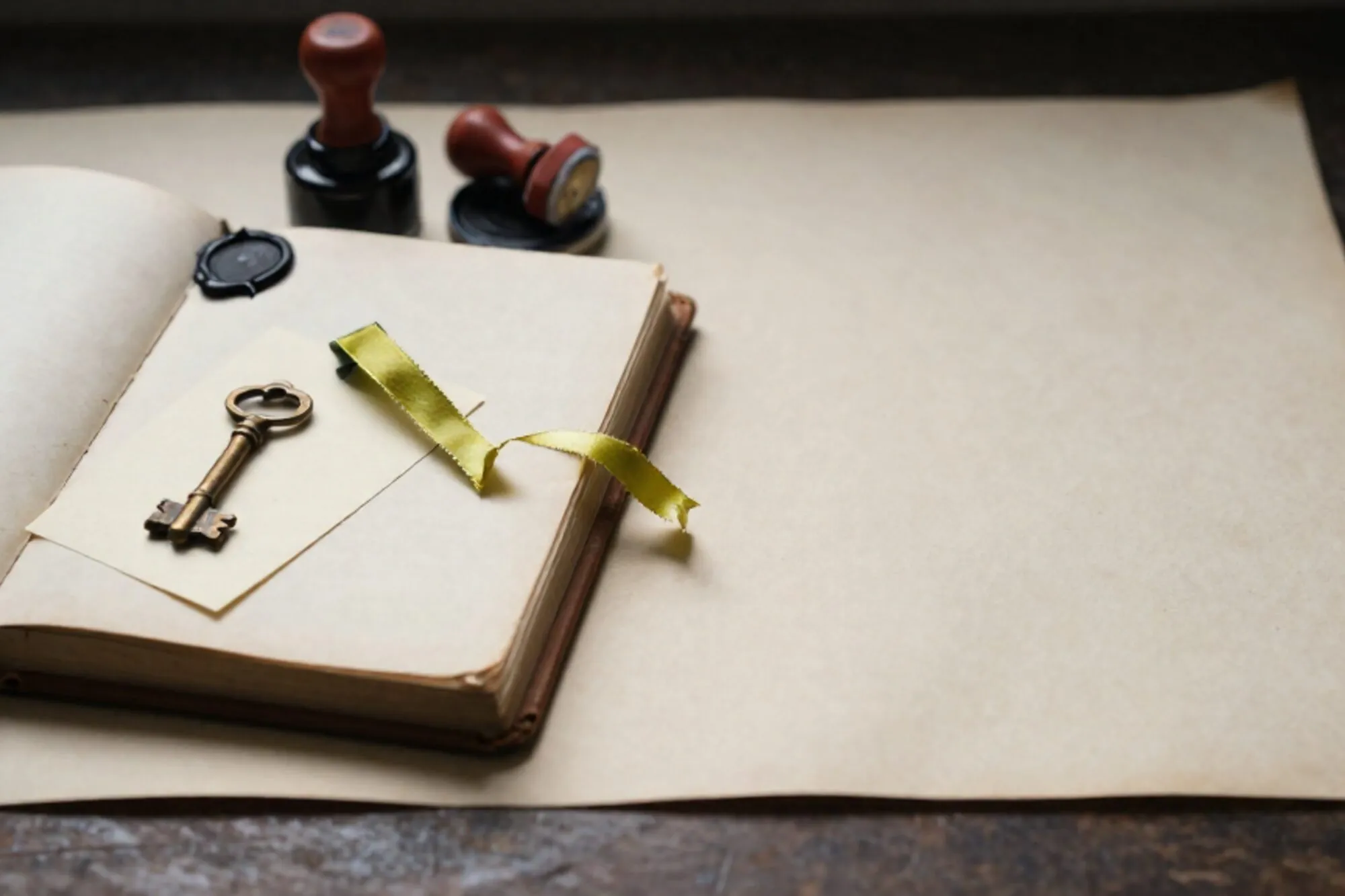 Open leather logbook with green ribbon, brass key on card, broken wax seal, red stamp, ink pad on ivory desk.