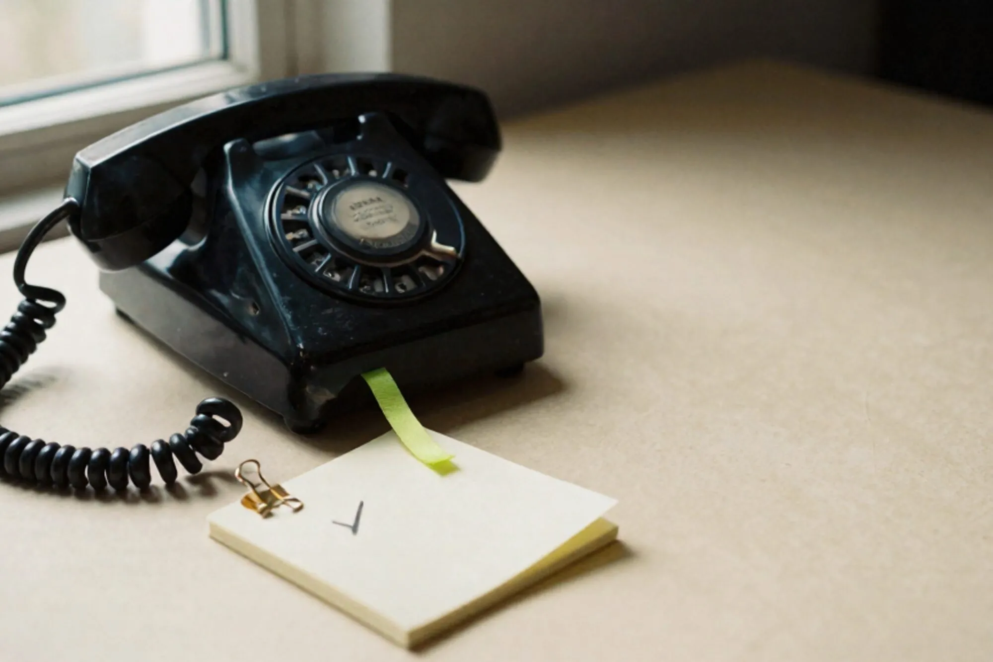 Black bakelite phone receiver off-hook on leather blotter, green ribbon bookmark, ivory notepad, brass paper clip.