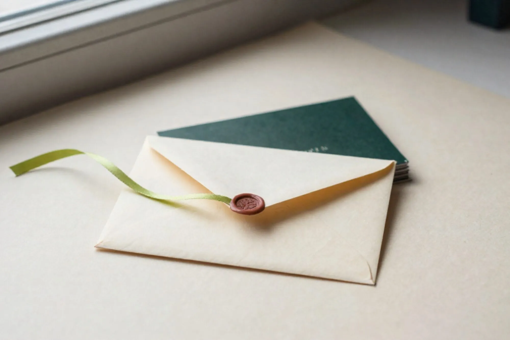 Cream envelope on ivory blotter, chartreuse ribbon across it, forest-green cards behind, red wax fragment, window light.