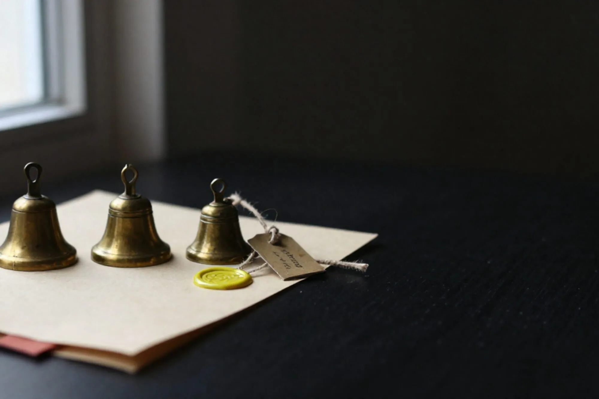 Three antique brass bells in a row on an ivory blotter, folded receipt with chartreuse wax seal, iron tag on twine.