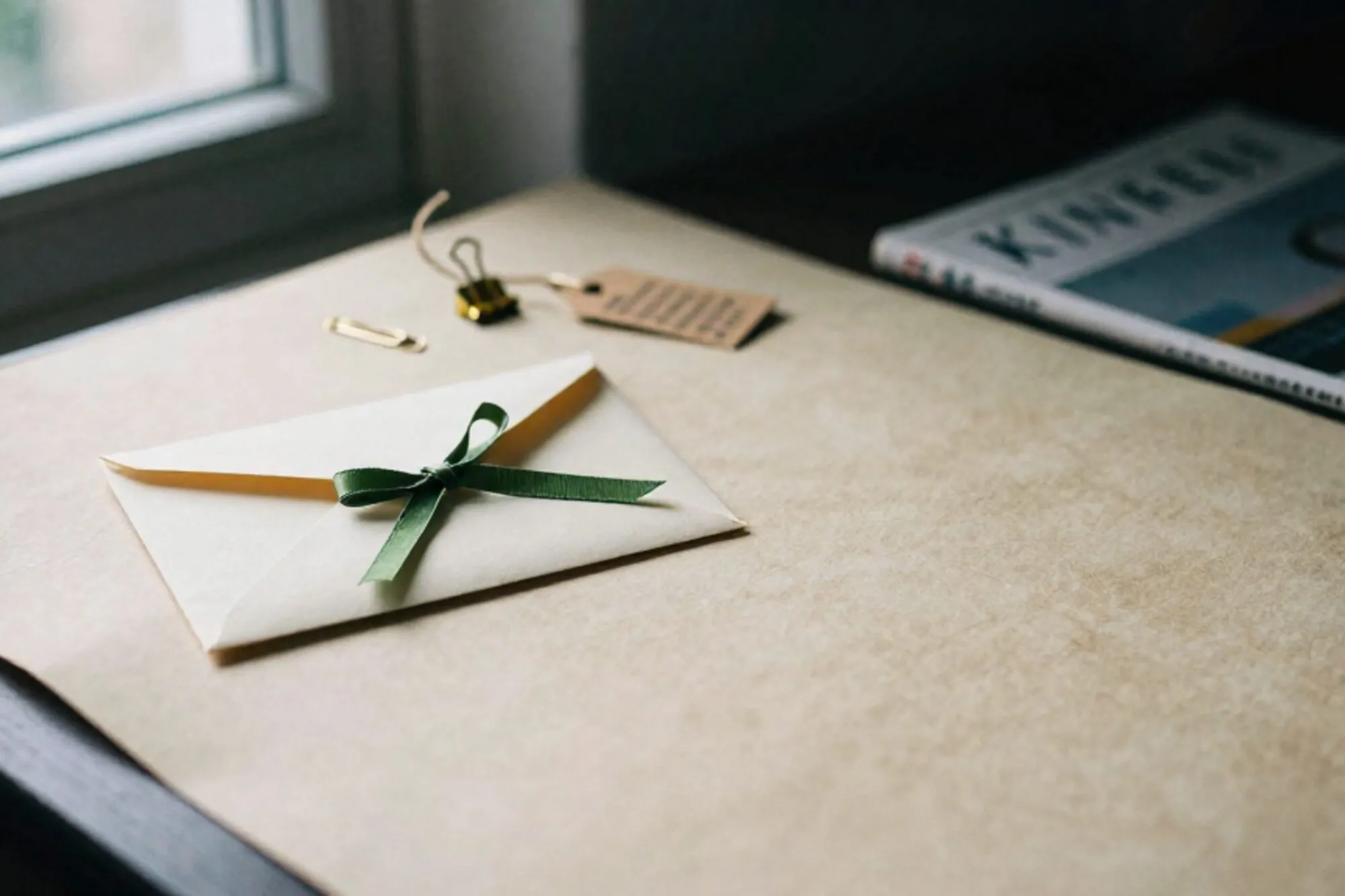 Cream envelope with chartreuse ribbon on ivory linen blotter, brass paper clip and shipping tag with twine beside it.