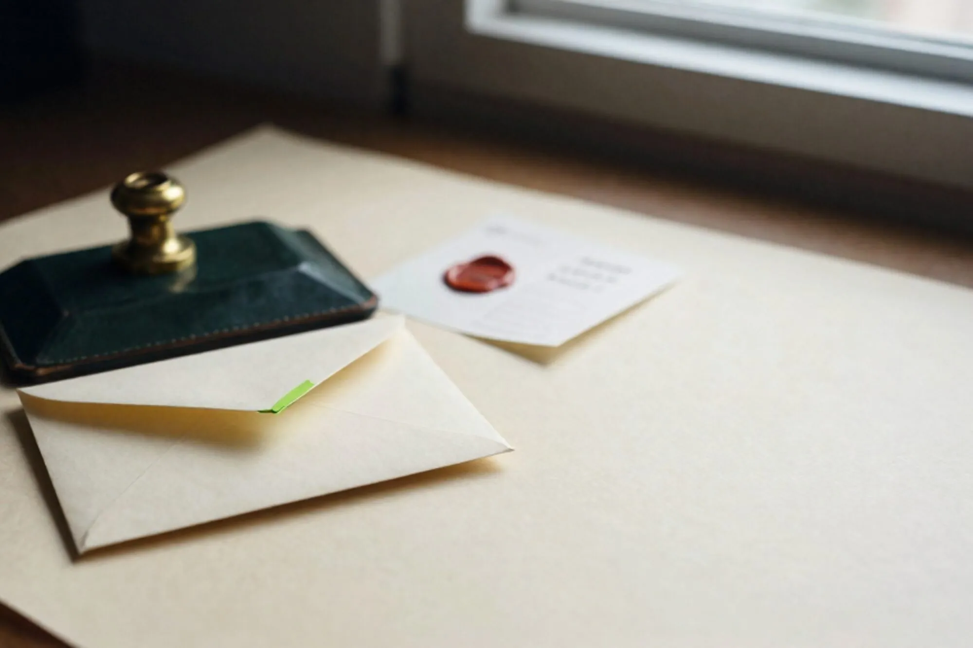 Crème envelop met chartreuse lint op donkergroene leren onderlegger, messing presse-papier, gevouwen memo, ivoren bureau.