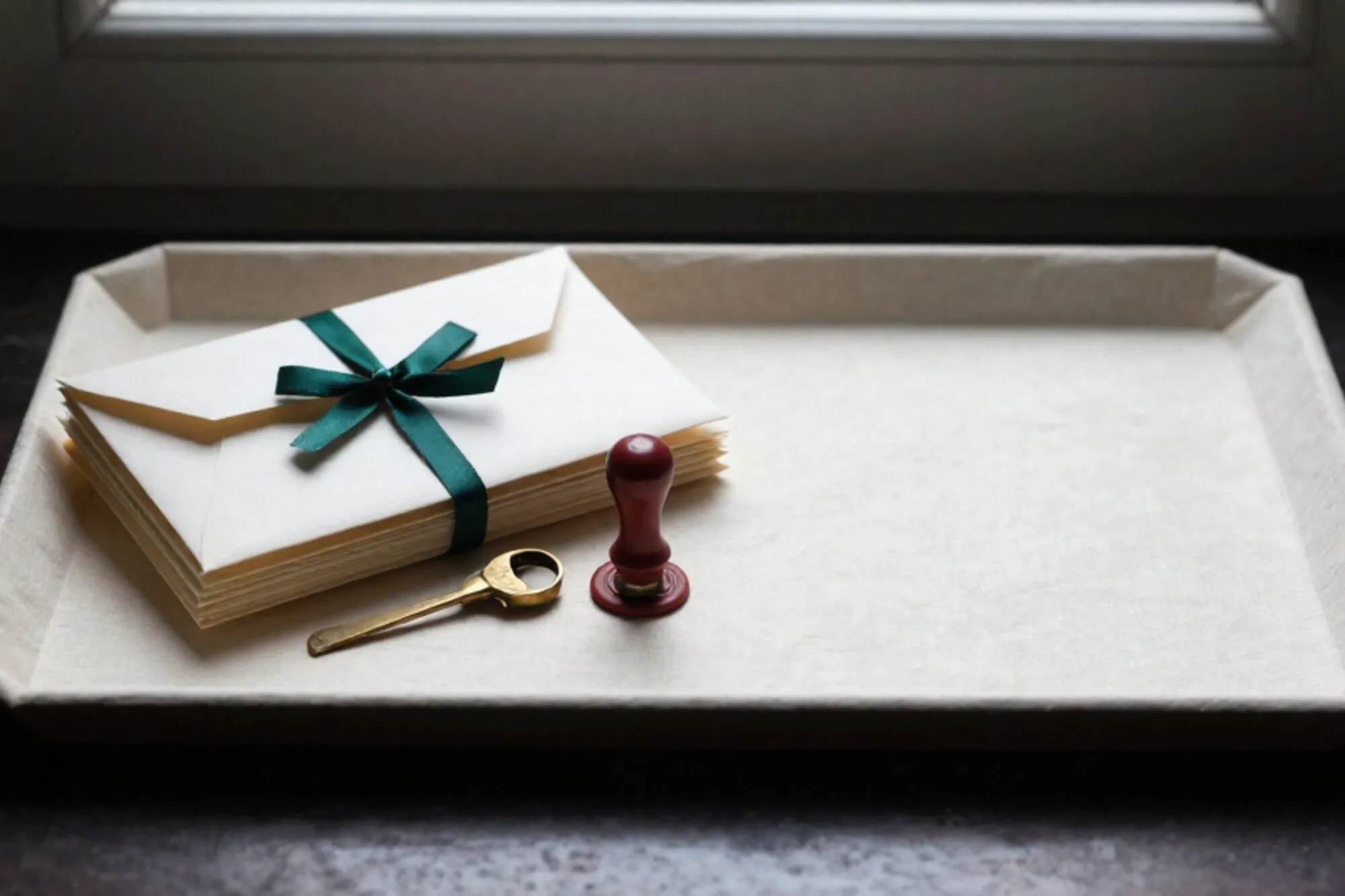 Linen tray with ivory envelopes tied in green ribbon, brass letter opener, red wax seal on bone-paper desk.