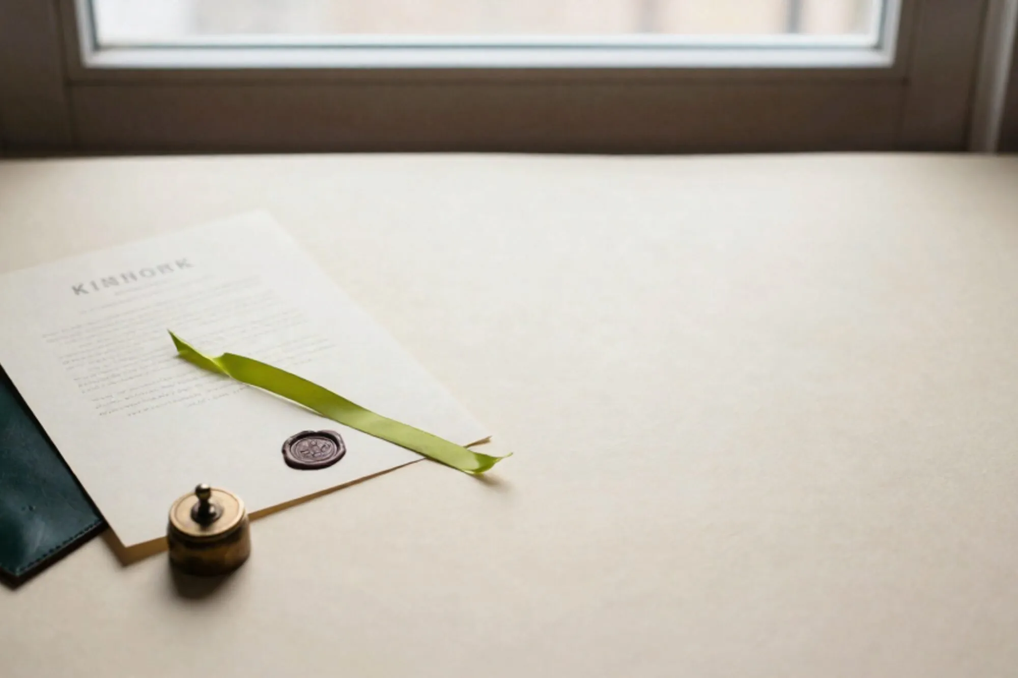 Cream parchment with wax seal, brass weight, green ribbon on ivory desk, forest leather blotter, side window light.