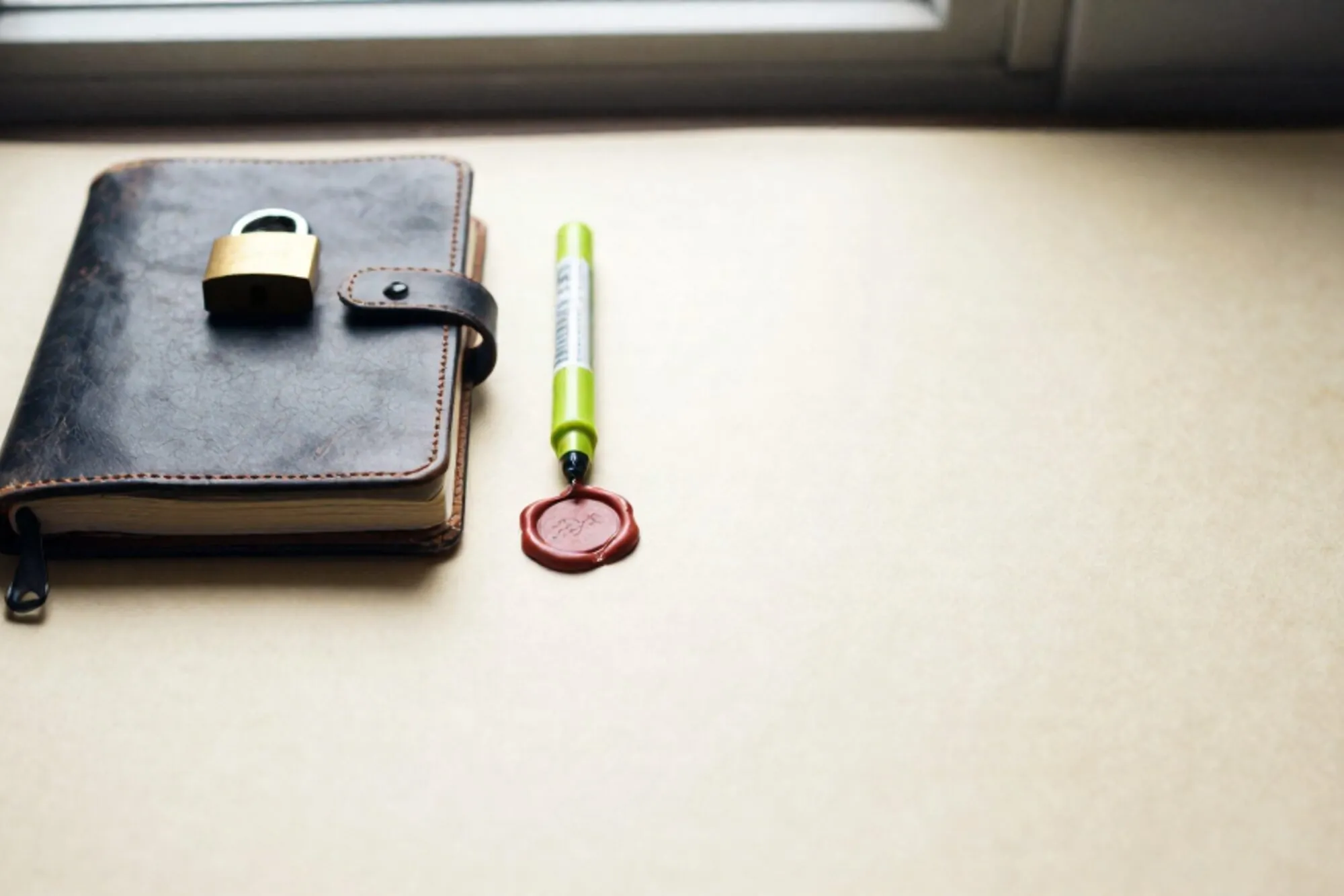 Closed leather ledger with brass padlock, green ribbon and cracked red wax seal on ivory paper.