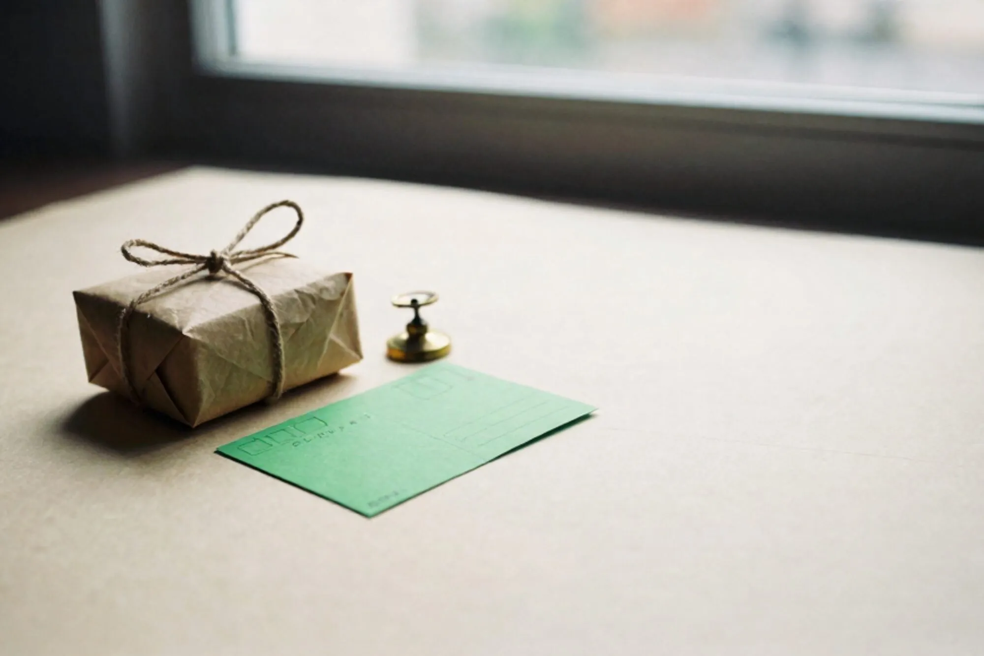Ivory paper surface with twine-tied craft parcel, folded chartreuse postcard, and small brass scale weight in side light.