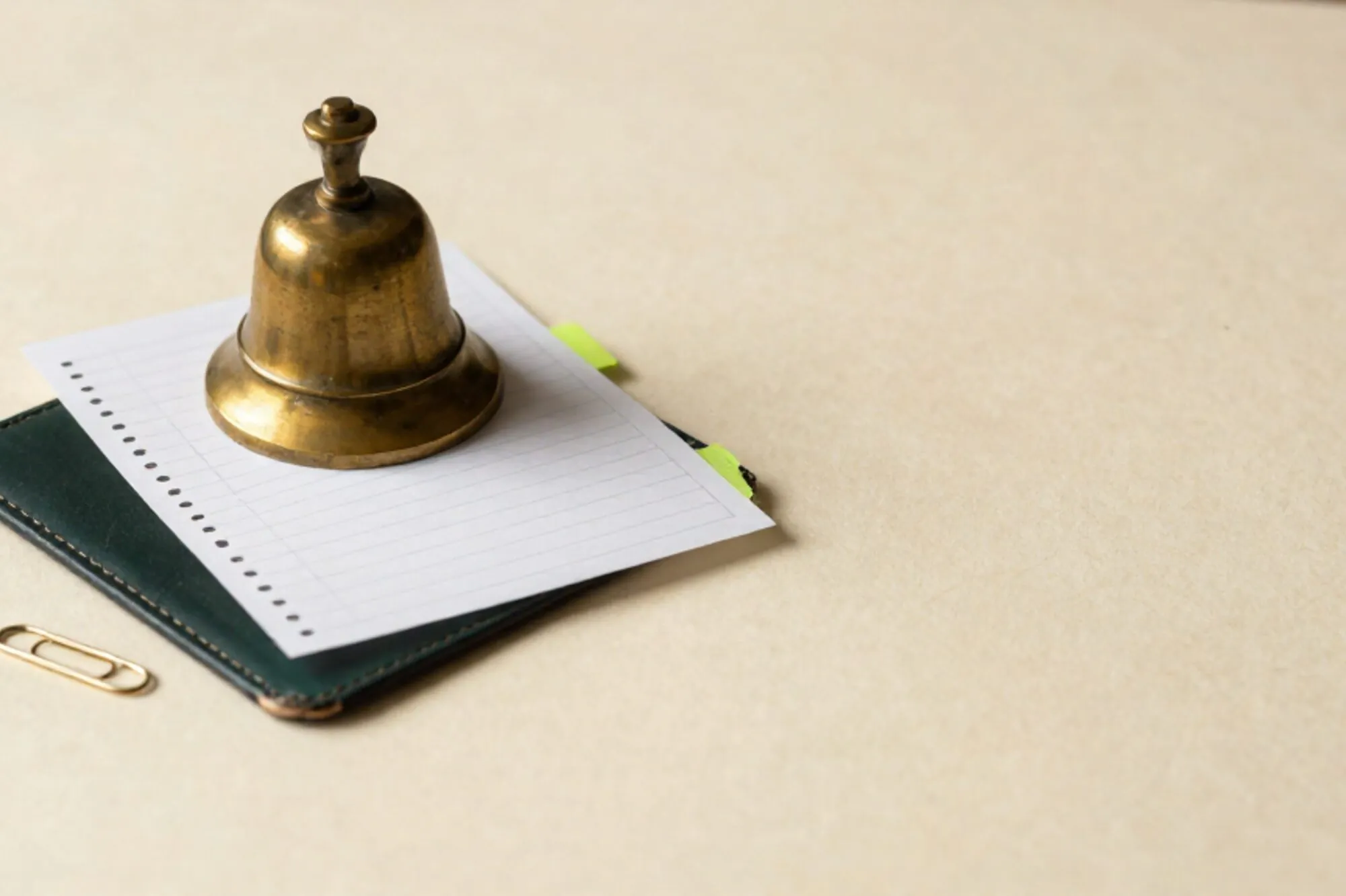Brass desk bell on ivory paper beside two stacked carbon-copy booking slips with a green sticky tab and paperclip.