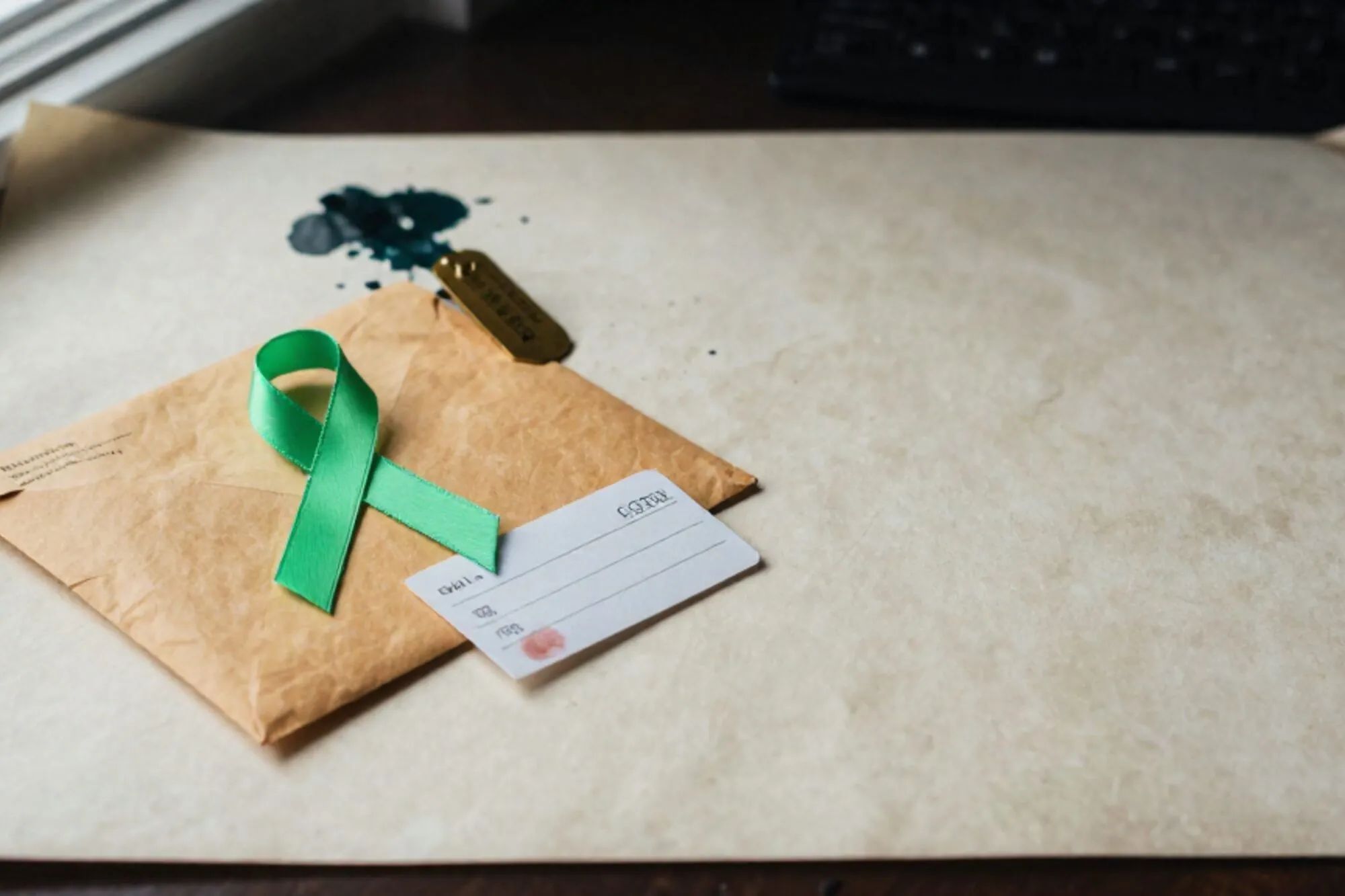 Manilla envelop met groen lint op ivoren bureau, koperen label en adreskaart ernaast, rode inktvlek, zijlicht.