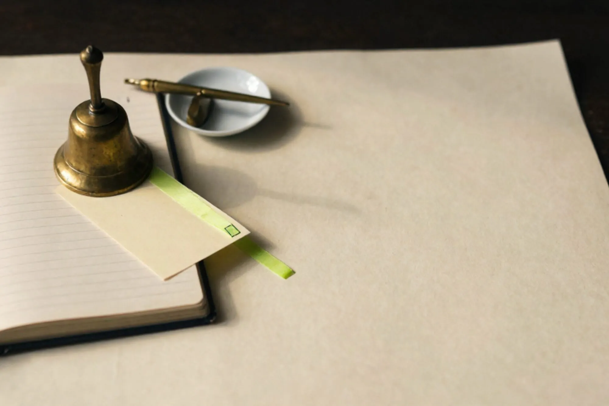 Brass call-bell on open linen ward ledger, folded card with chartreuse ribbon, porcelain saucer with pen rest.