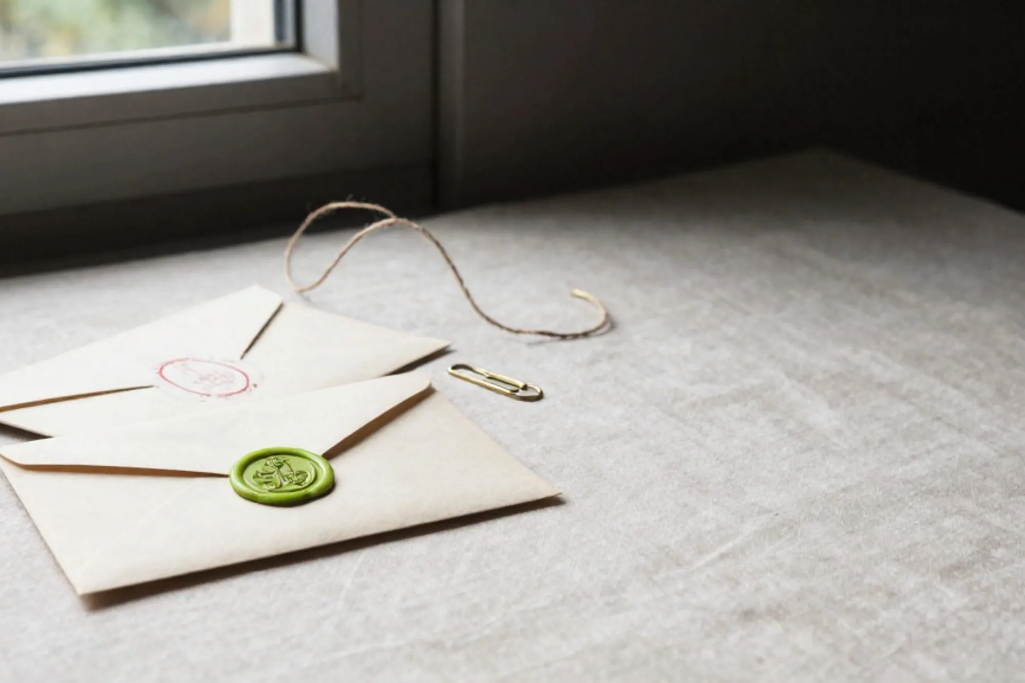 Twee crème enveloppen met rode stempels en een groene lakzegel, messing paperclip en jute touw op linnen.