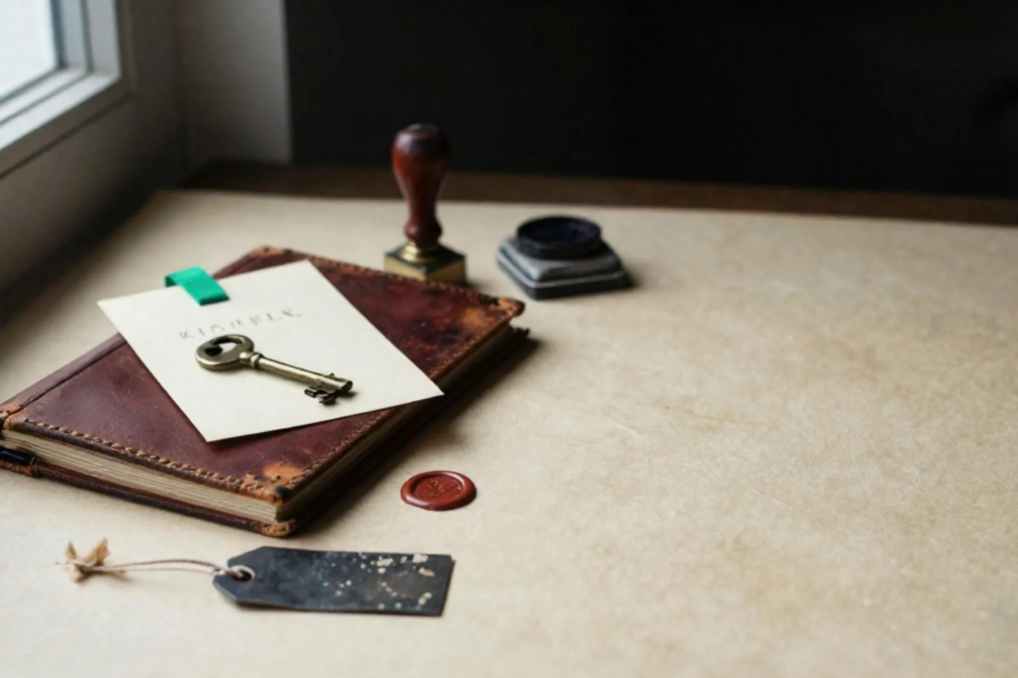 Leather logbook, brass key on cream card with green ribbon, wax seal fragment and shipping tags on ivory desk.