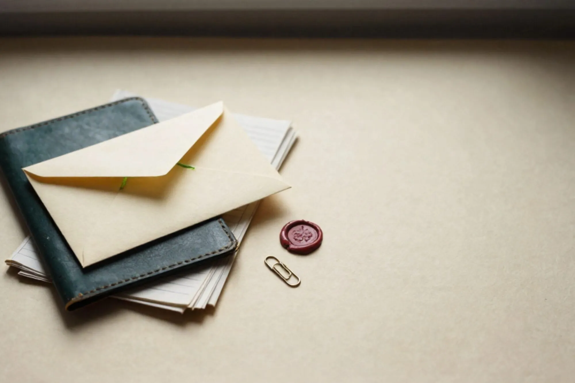 Crèmekleurige envelop met limoengroen lint op donkergroene leren onderlegger, grootboekpapier, koperen paperclip, rode lakzegel.