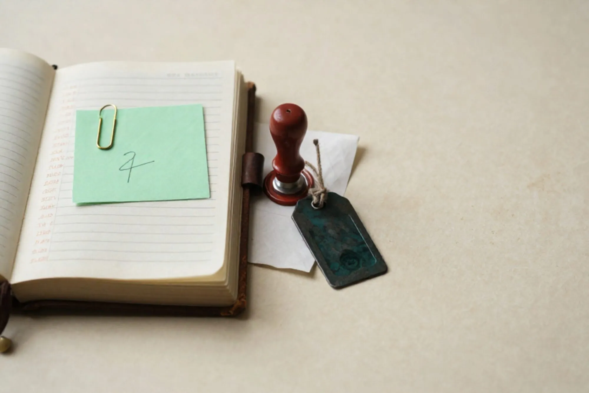 Open leather ledger with copperplate entries, brass paperclip on green index card, red wax seal, iron luggage tag.