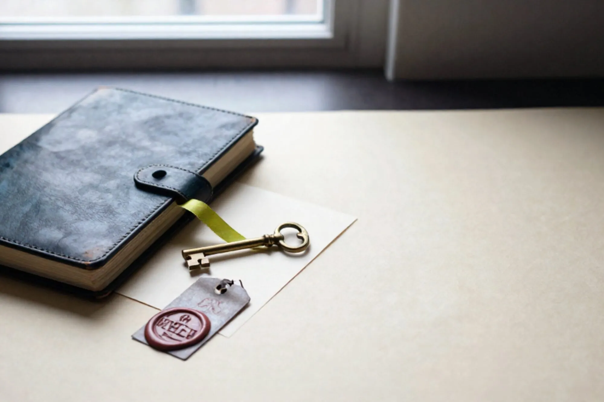 Open leather ledger with brass key, cream card, green silk ribbon, and red wax seal on ivory paper by a window.