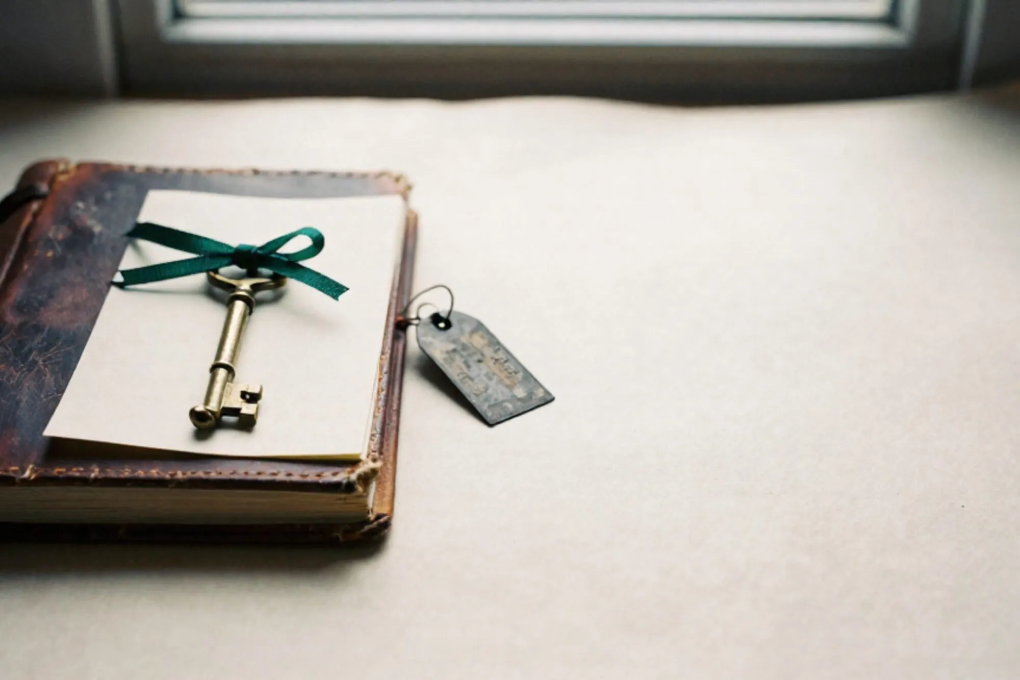 Leather logbook, brass key on cream card with green ribbon, iron tag on ivory paper, side window light.
