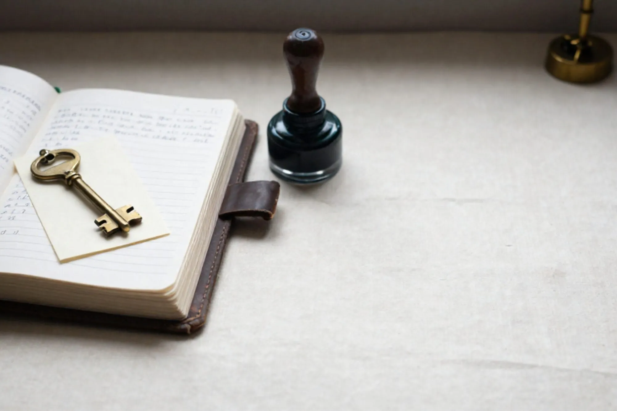 Open leather logbook with handwritten pages, brass key on cream card, rubber stamp, green ribbon on ivory linen.