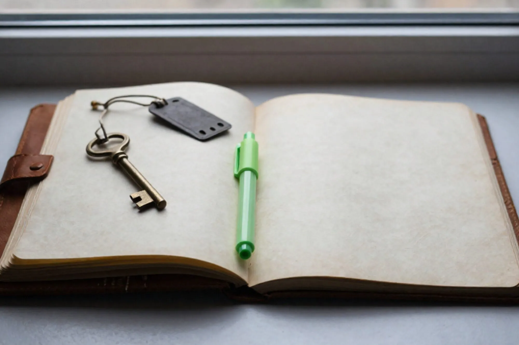 Open leather logbook with brass key, green ribbon marker, iron shipping tag on ivory blotter by window.