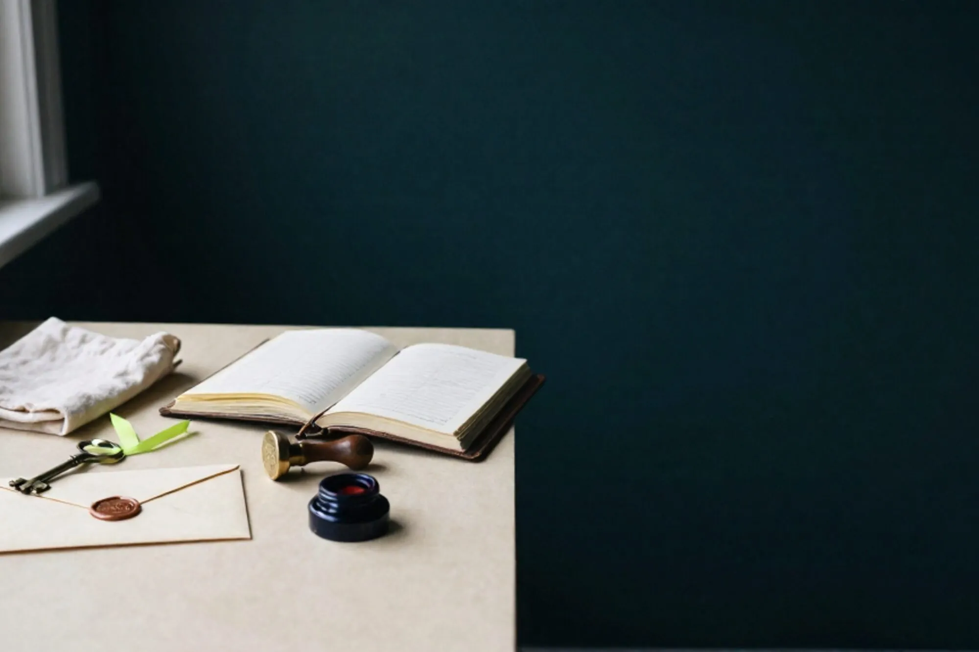 Brass key with green ribbon on linen, wax-sealed envelope, leather ledger, rubber stamp and ink pad on ivory desk.
