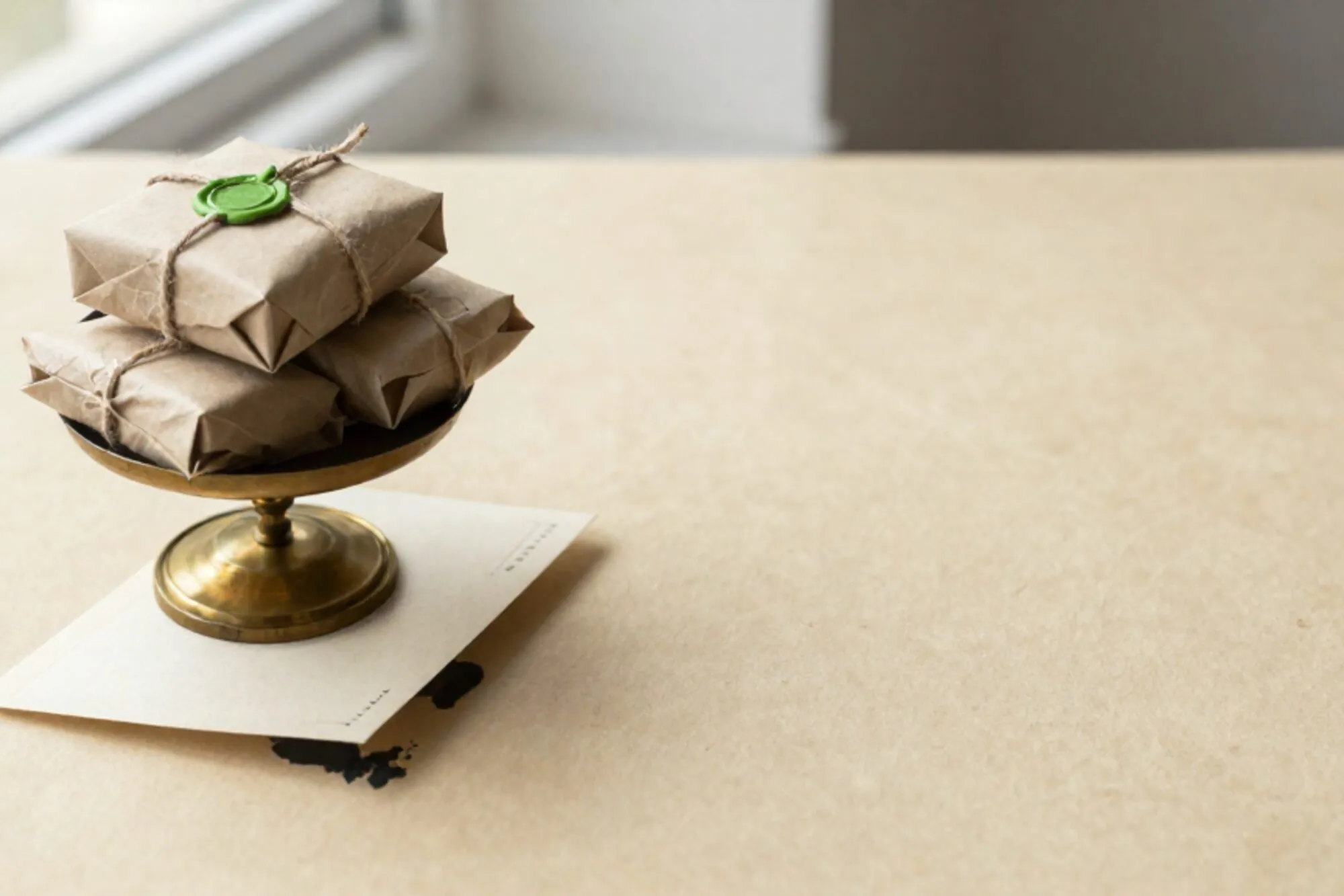 Brass weighing scale holding three twine-tied craft paper parcels on ivory paper, cream postcard tucked beneath.