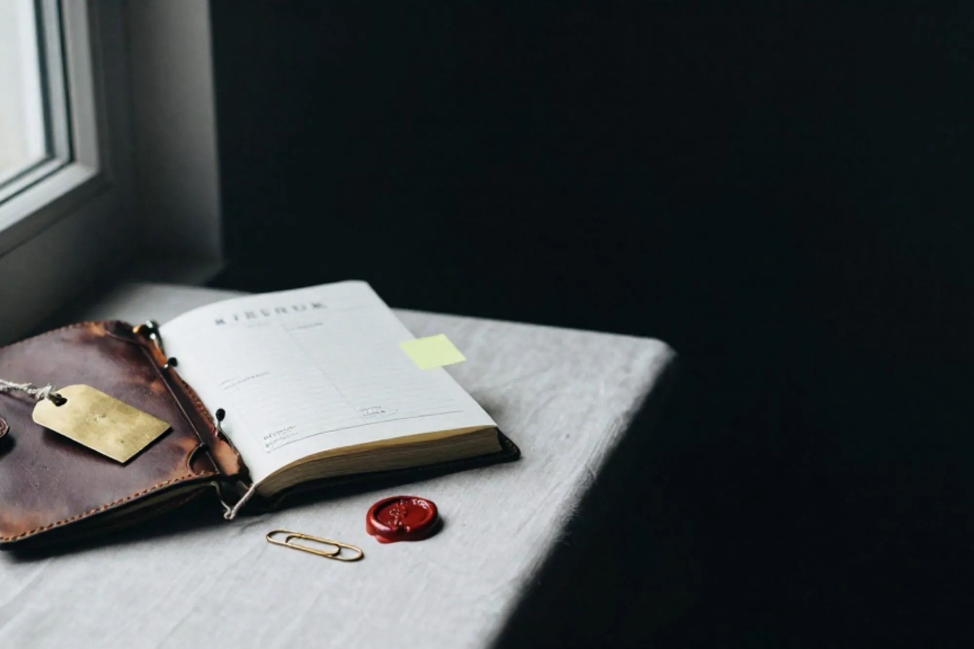 Half-open leather ledger with brass tag, carbon invoice, green sticky tab and wax stub on ivory linen, dark green backdrop.