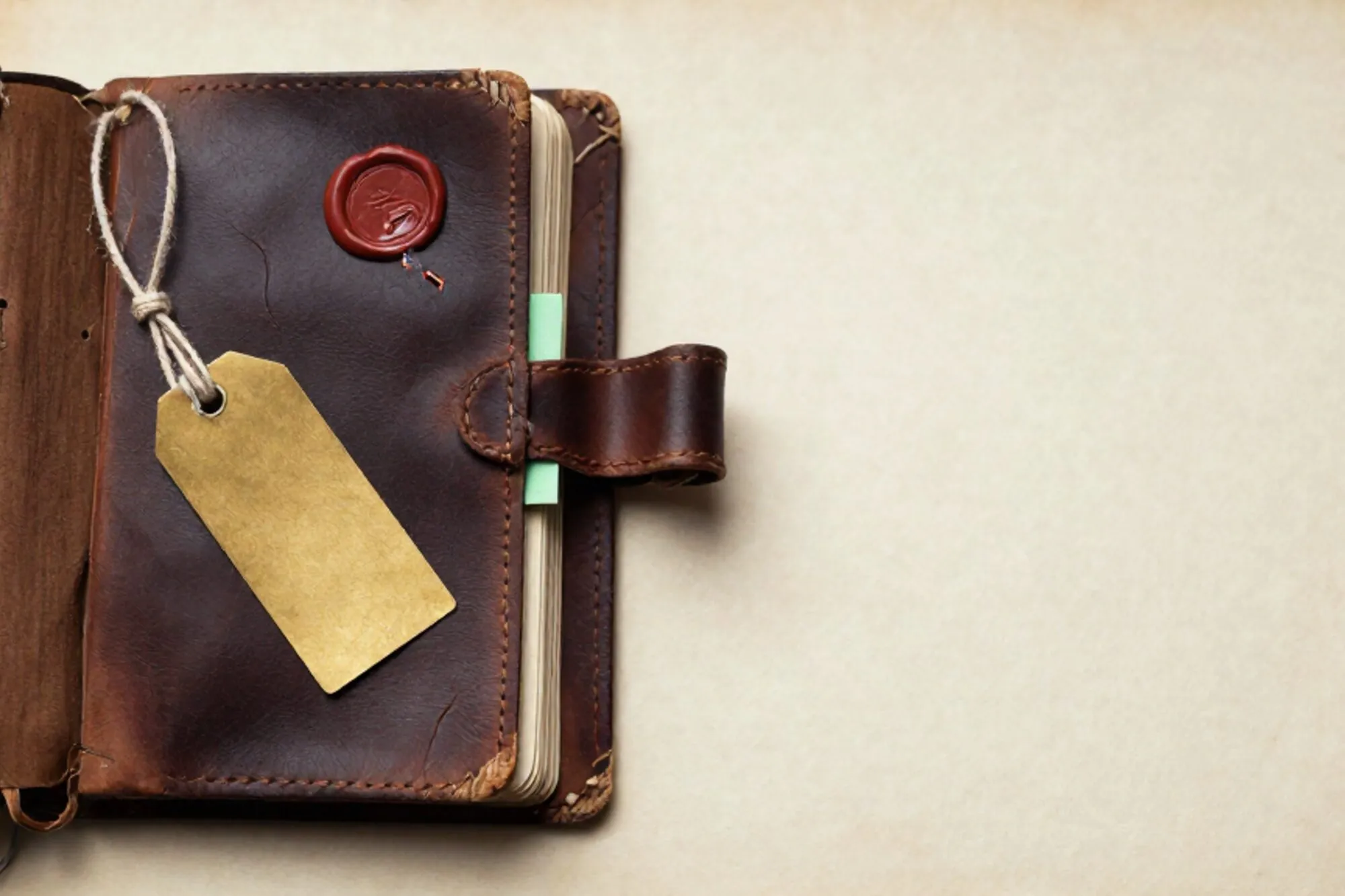 Worn leather ledger half-open on ivory paper, brass shipping tag on linen twine, green index card, broken red wax seal.