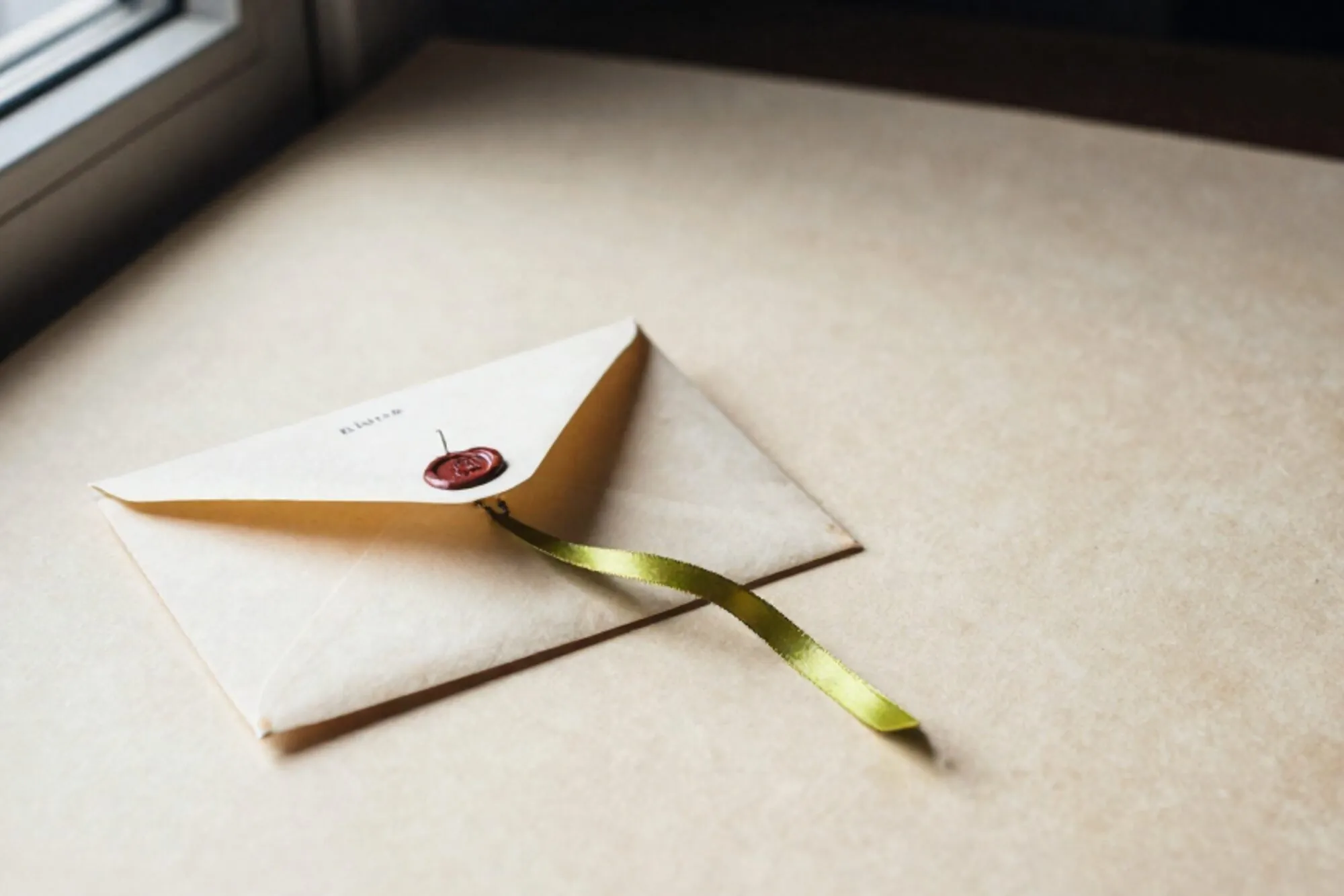 Manila envelope on linen blotter, chartreuse silk ribbon, brass tag, red wax seal, ivory paper, forest shadows.