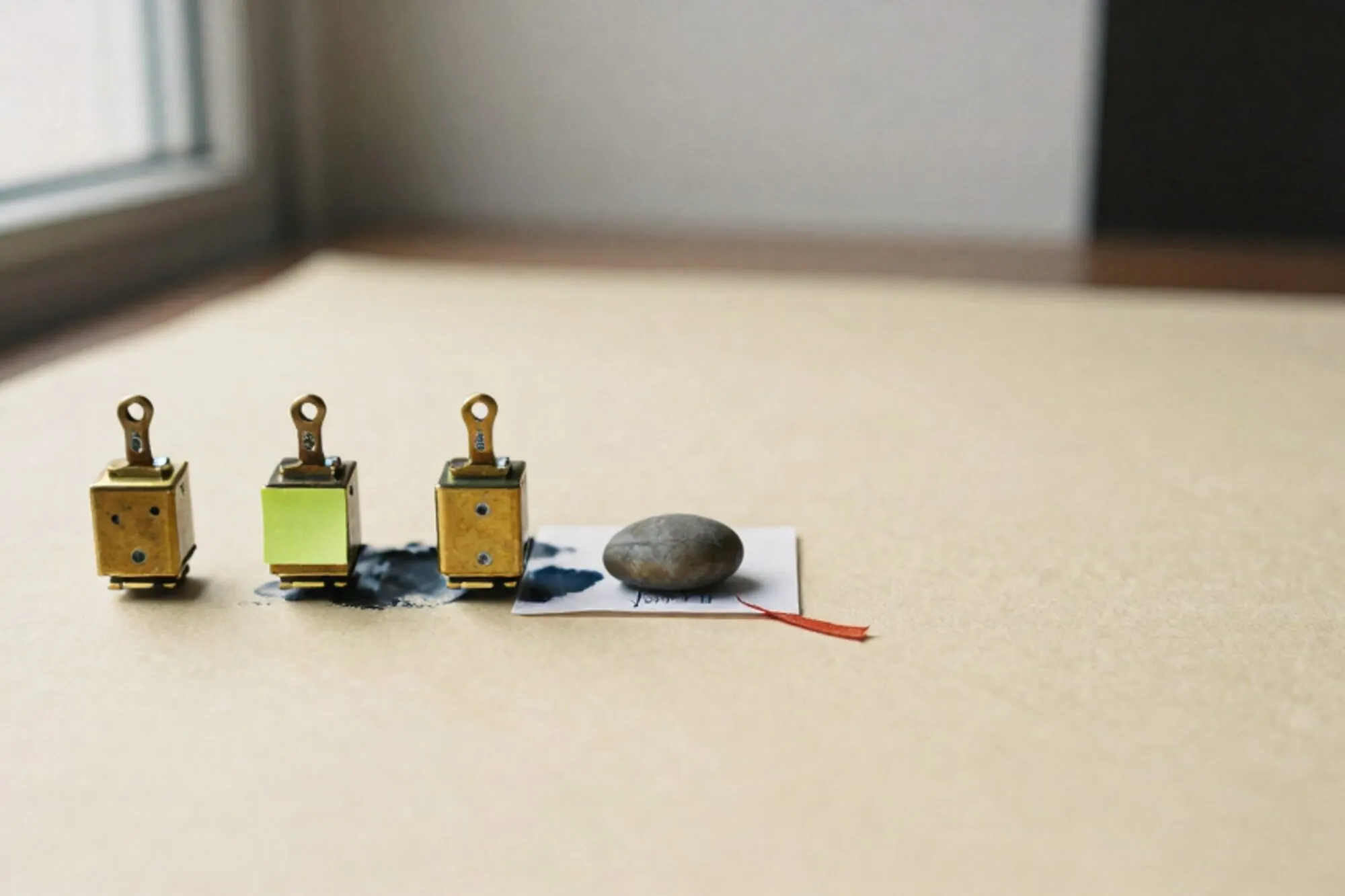 Three brass relays in a row on ivory paper, folded form with river stone, green sticky note, red ribbon, soft shadows.