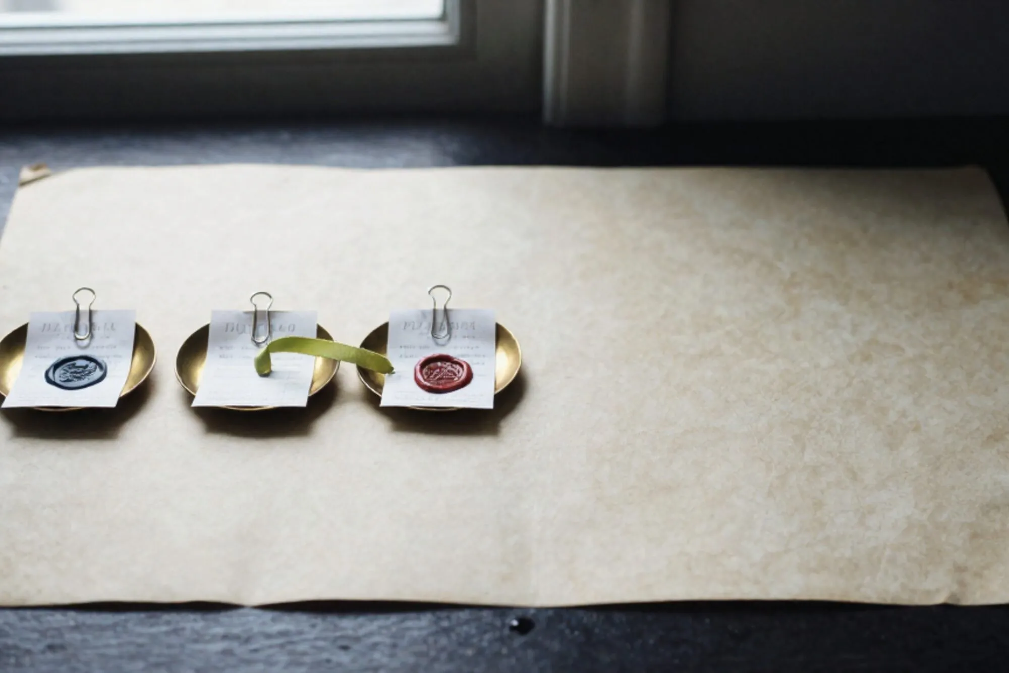 Three brass dishes on ivory linen hold folded receipts with wax seals; a green ribbon threads the middle dish.