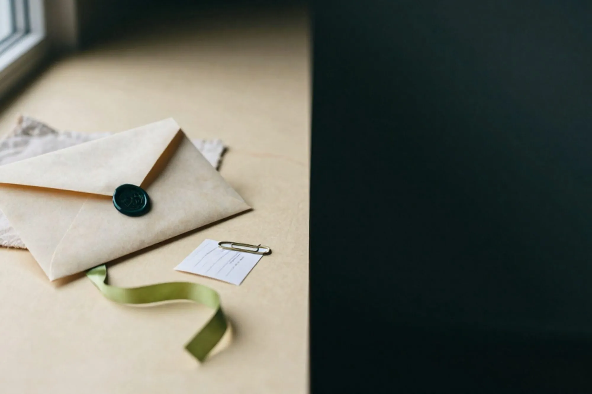 Crème envelop met waszegel op linnen onderlegger, lichtgroen zijden lint, koperen paperclip, gevouwen doorslagbon op ivoor.