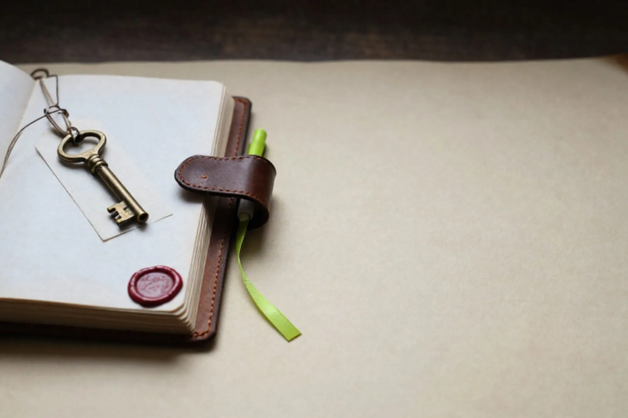 Open leather logbook on ivory paper desk, brass key on tag with twine, green ribbon, red wax seal fragment.