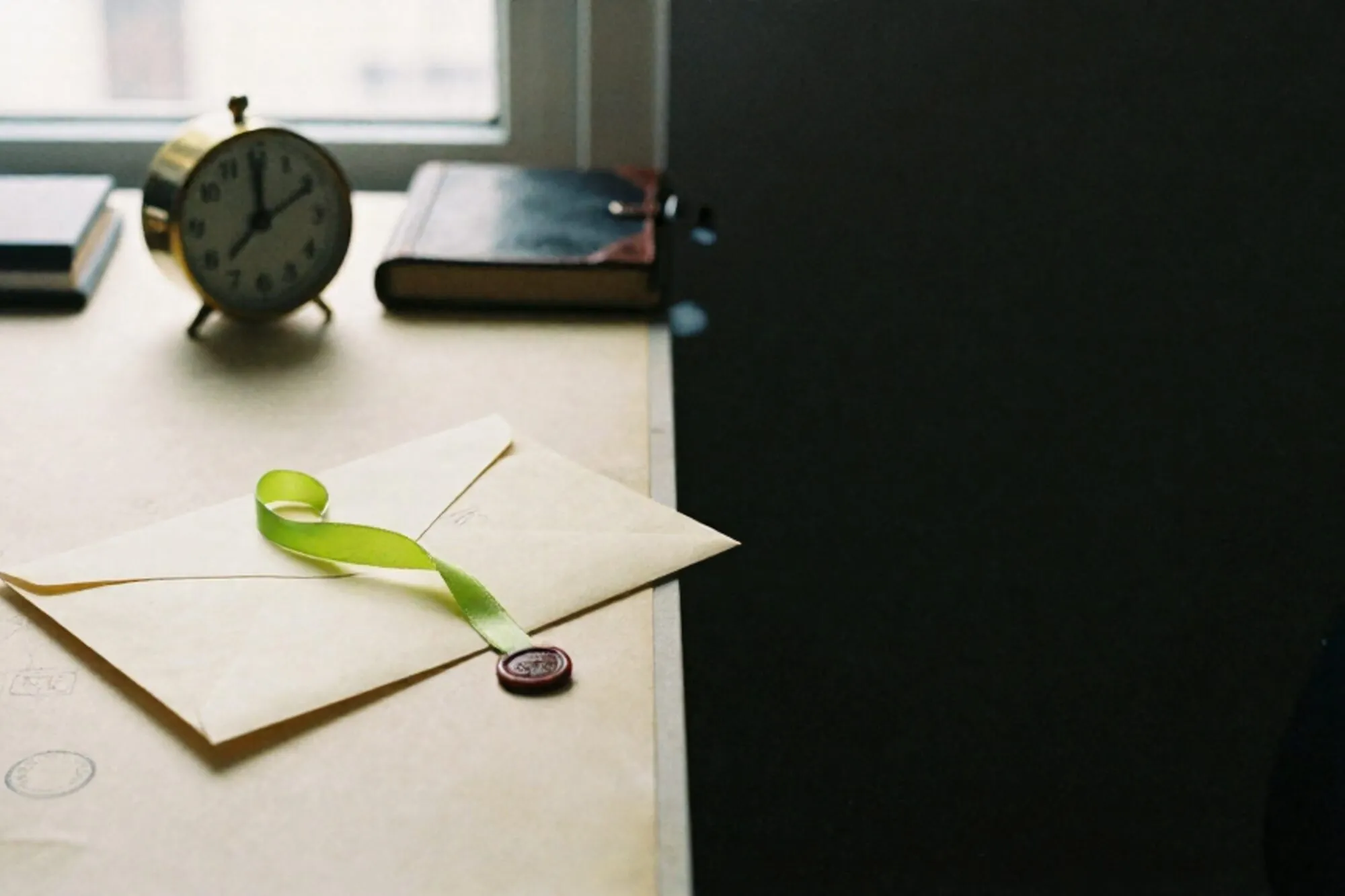 Cream envelope with chartreuse ribbon, brass clock at 15:48, leather logbook, red wax fragment on ivory desk blotter.