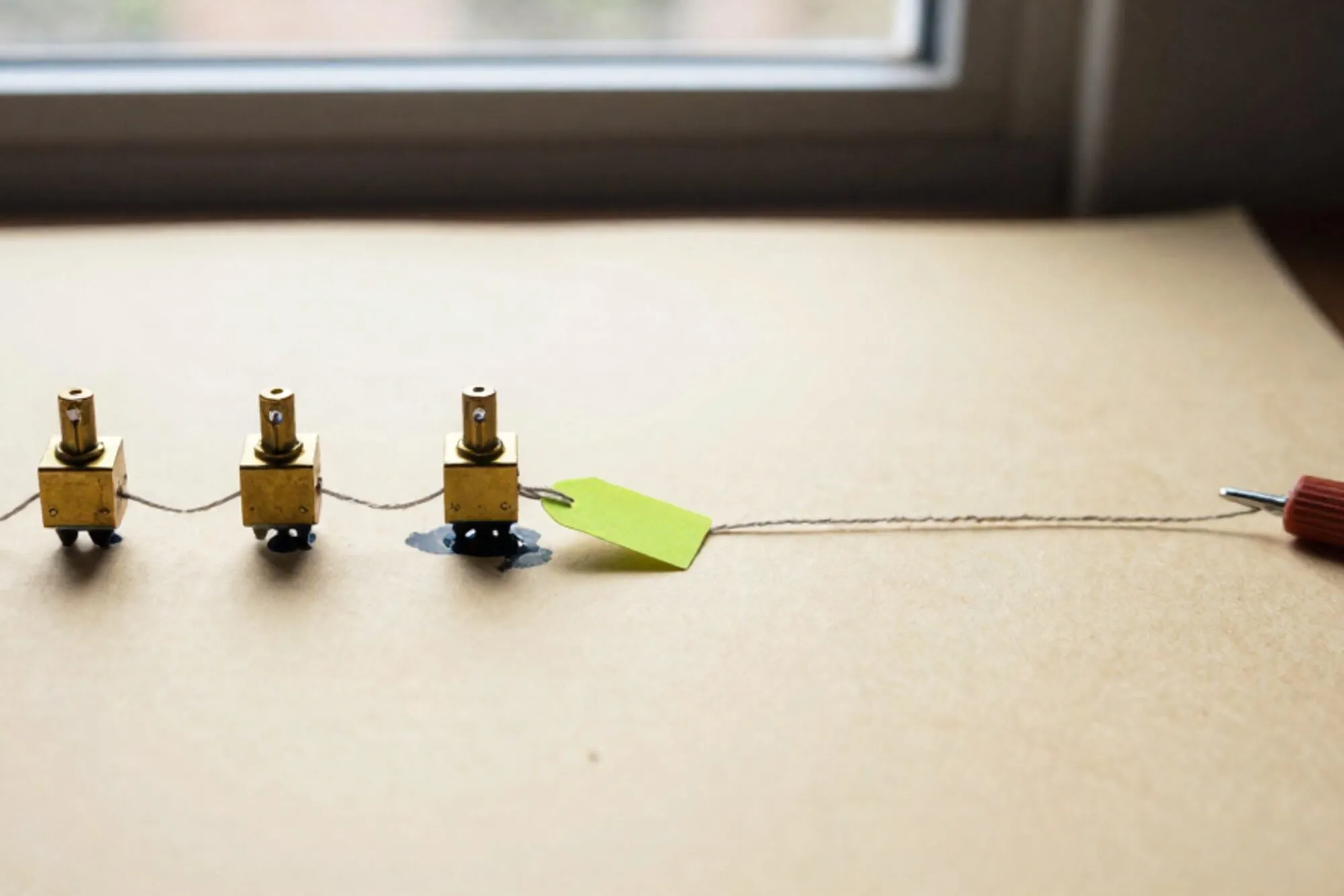 Three brass relay switches in a row linked by waxed thread on ivory paper, green tag on middle relay, red ribbon at edge.